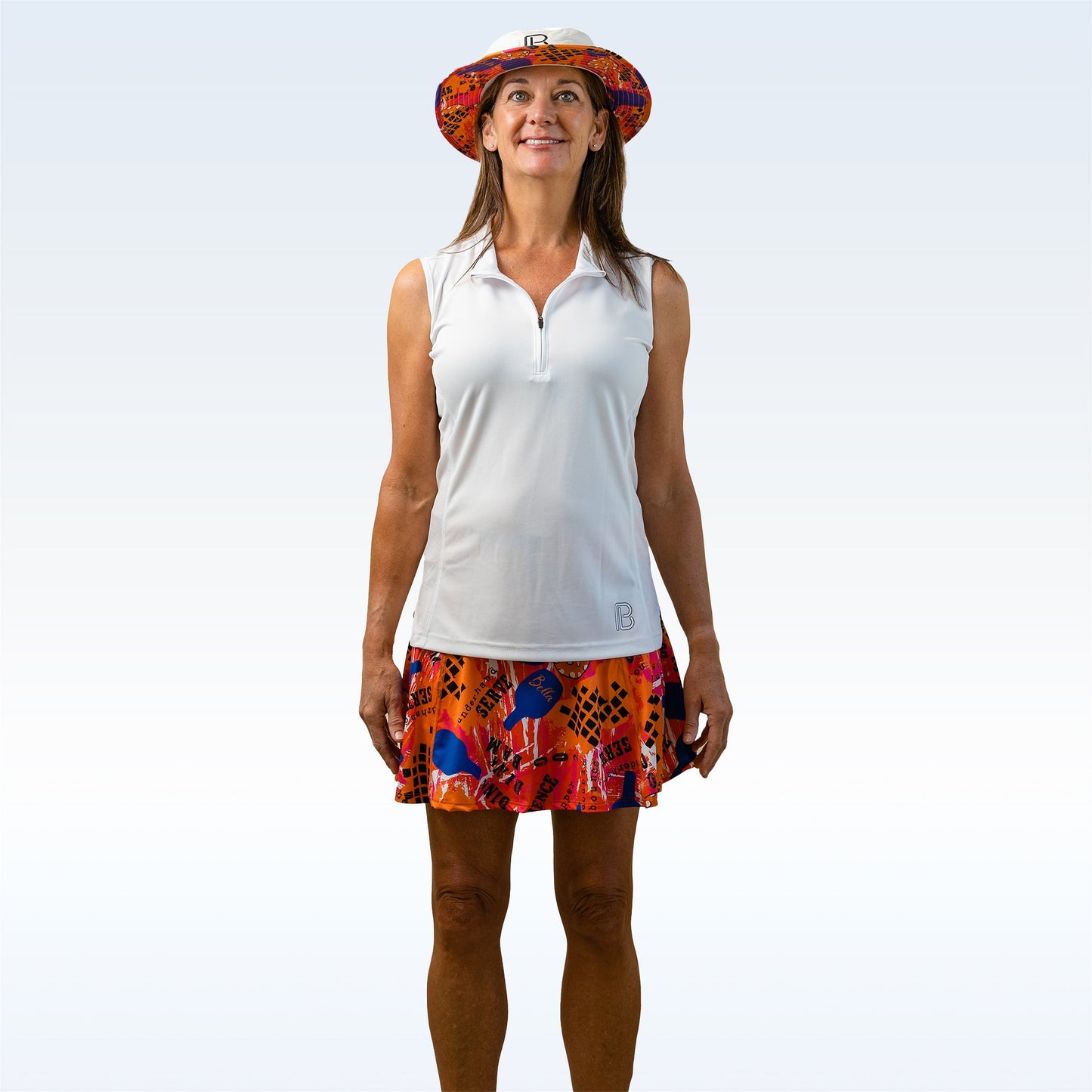 A woman smiles against a light background, wearing the Pickleball Bella PB 1/4 Zip Sleeveless White top and a red-blue patterned stretch skirt. She faces forward in a wide-brimmed hat that provides SPF 50+ UV protection.
