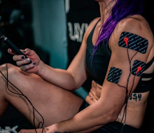 A muscular person with purple hair, black nails, and dressed in a sports bra and shorts uses the iReliev SPORT Large Pads Refill Kit, attaching snap style electrode pads to their upper arm and forearm for targeted muscle stimulation.