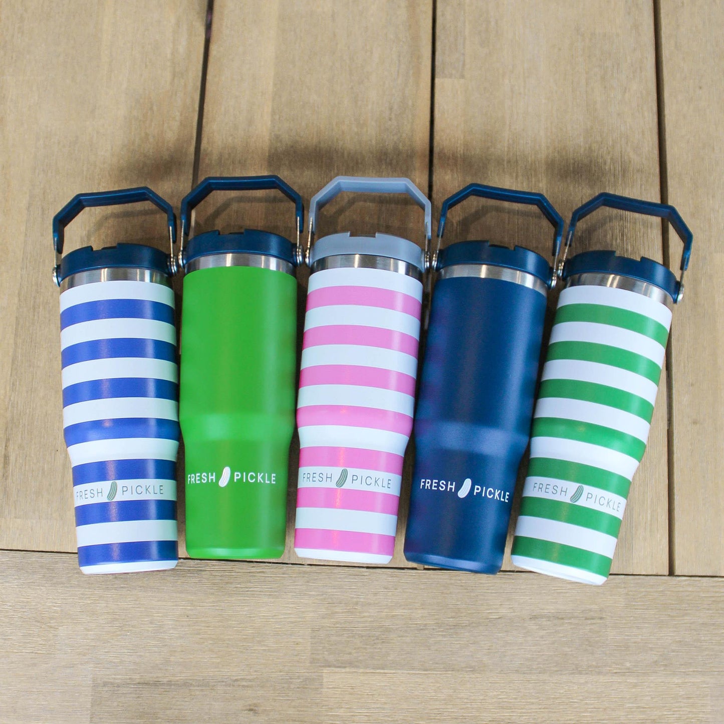 Five Fresh Pickle Tumbler cups with handles sit on a wooden surface. Each 30 oz, double-walled, vacuum-sealed tumbler comes in blue stripes, solid green, pink stripes, solid blue, and green stripes—all labeled Fresh Pickle.