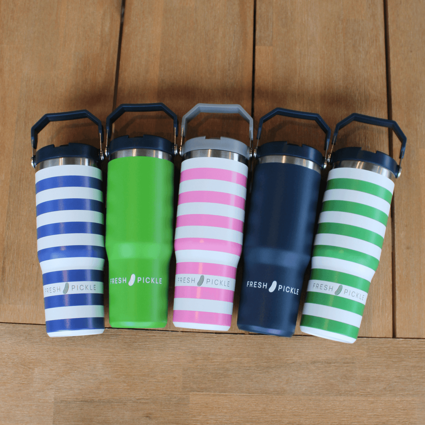 Five Fresh Pickle Tumblers by Fresh Pickle, including a 30 oz water bottle, are displayed on wood. Designs feature blue/white stripes, solid green, pink/white stripes, solid navy blue, and green/white stripes—each with a handle.
