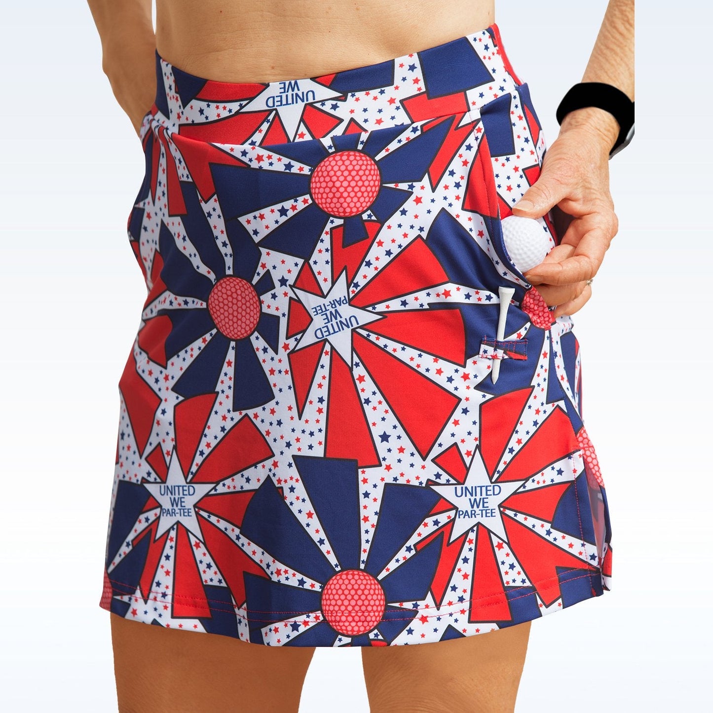 A person wears the Pickleball Bella United We Par-Tee Straight Golf Skort featuring red, white, and blue star patterns and "UNITED WE PARTEE" text, holding a golf ball and sporting a black wristband against a light background.