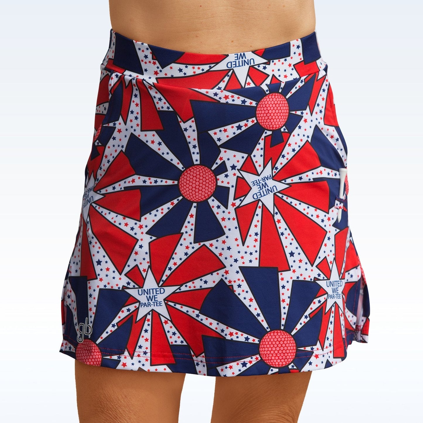 Someone wears the Pickleball Bella United We Par-Tee Straight Golf Skort, featuring a red, white, and blue geometric pattern with starbursts, dots, and "UNITED WE PARTY" text. The above-the-knee hem sits on a pale gradient background.