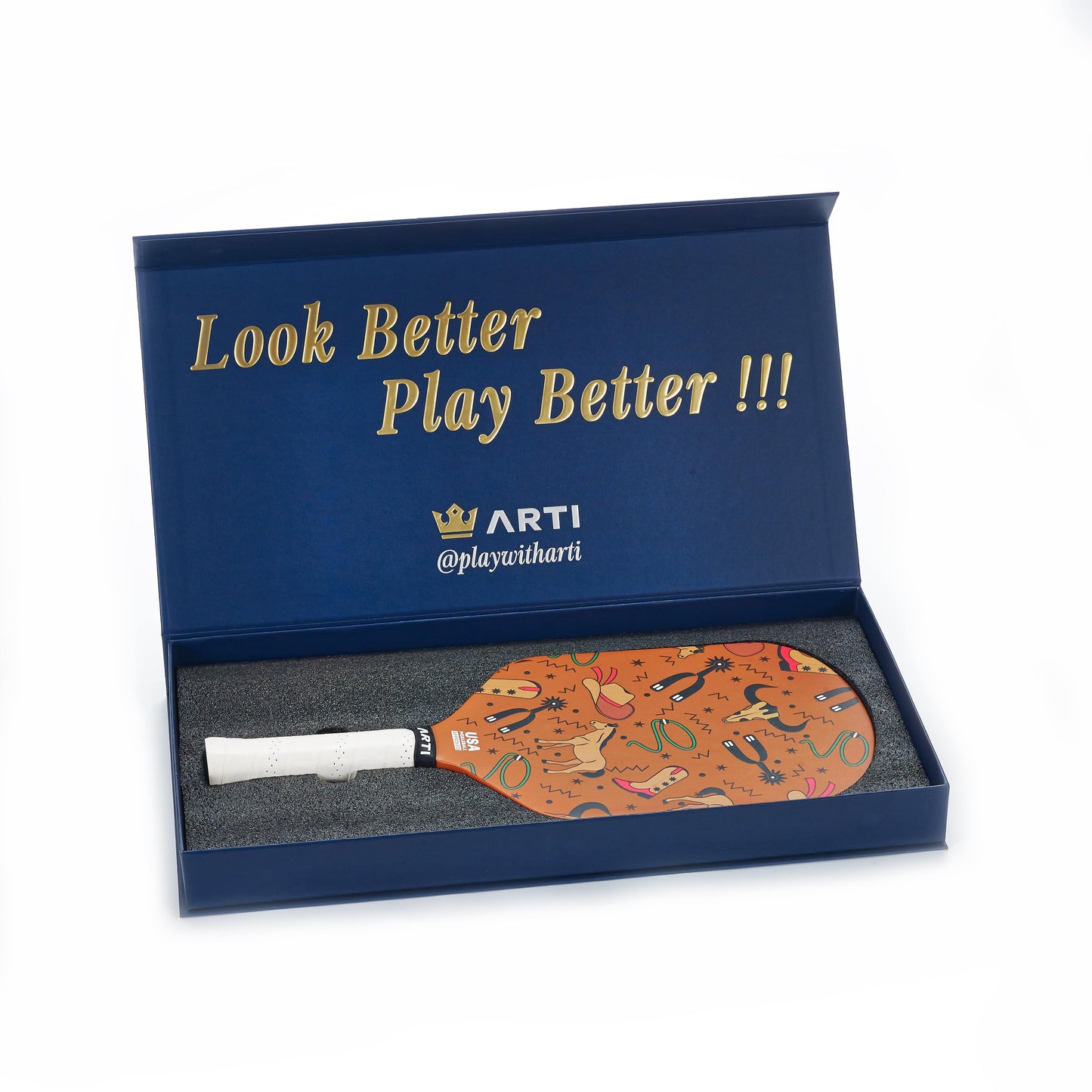 A blue ARTI box with gold “Look Better Play Better !!!” and “ARTI @playwitharti” holds the USAPA-approved ARTI Carbon Fiber "TEXAS" Paddle, featuring playful patterns, a white handle, and resting in black foam inside the open box.