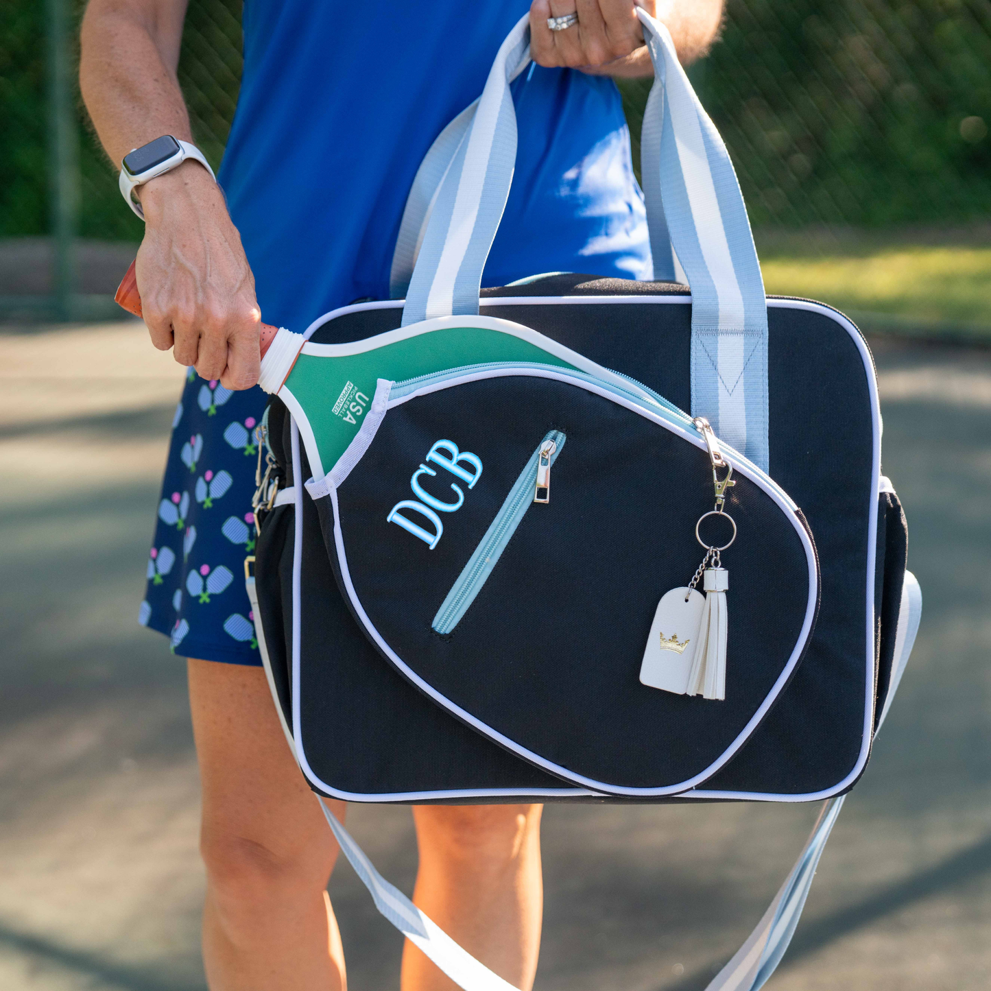 A person in a blue skirt with a tennis racquet print carries the Queen of the Court Prepster Pickleball Bag, monogrammed “DCB” and holding a green racquet. The bag, ideal for pickleball gear, features white-tag keychains on a sunlit tennis court.