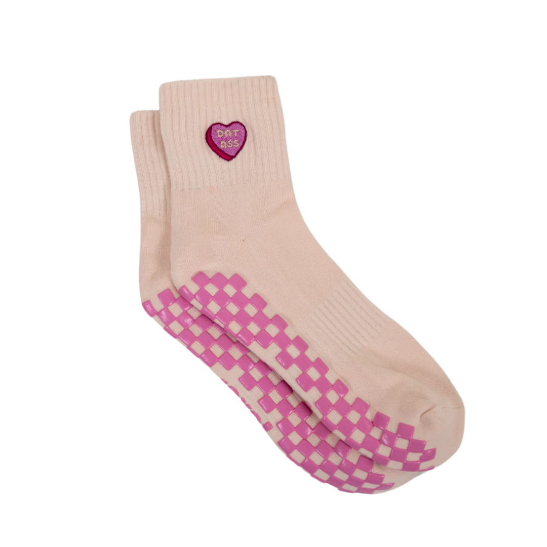 The Sweet Nothings Grip Sock by Revel features a light pink hue with pink and purple checkerboard grips and a small embroidered heart patch near the top that reads "DONT ASS," shown neatly stacked on a plain white background.