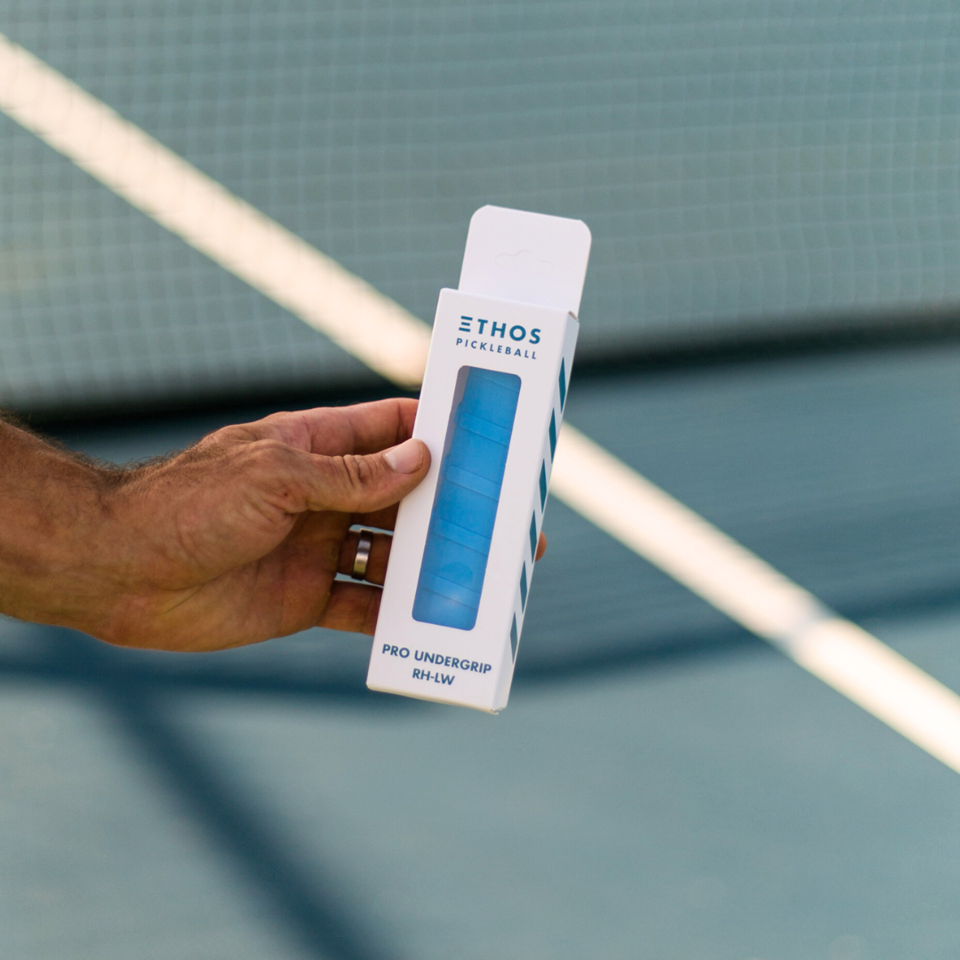 A hand holds an Ethos Pickleball Pro Undergrip RH-LW—Right Hand Specific, Lightweight, Vibration-Reducing Grip—in blue, shown through white and blue packaging in front of a pickleball court net.
