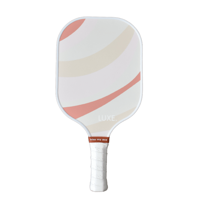 The Pink and Rust paddle by LUXE Pickleball features a white face with abstract pink and beige stripes, "LUXE." printed near the bottom, a textured white grip, and a red band with "kiss my ace" at the top of the handle.
