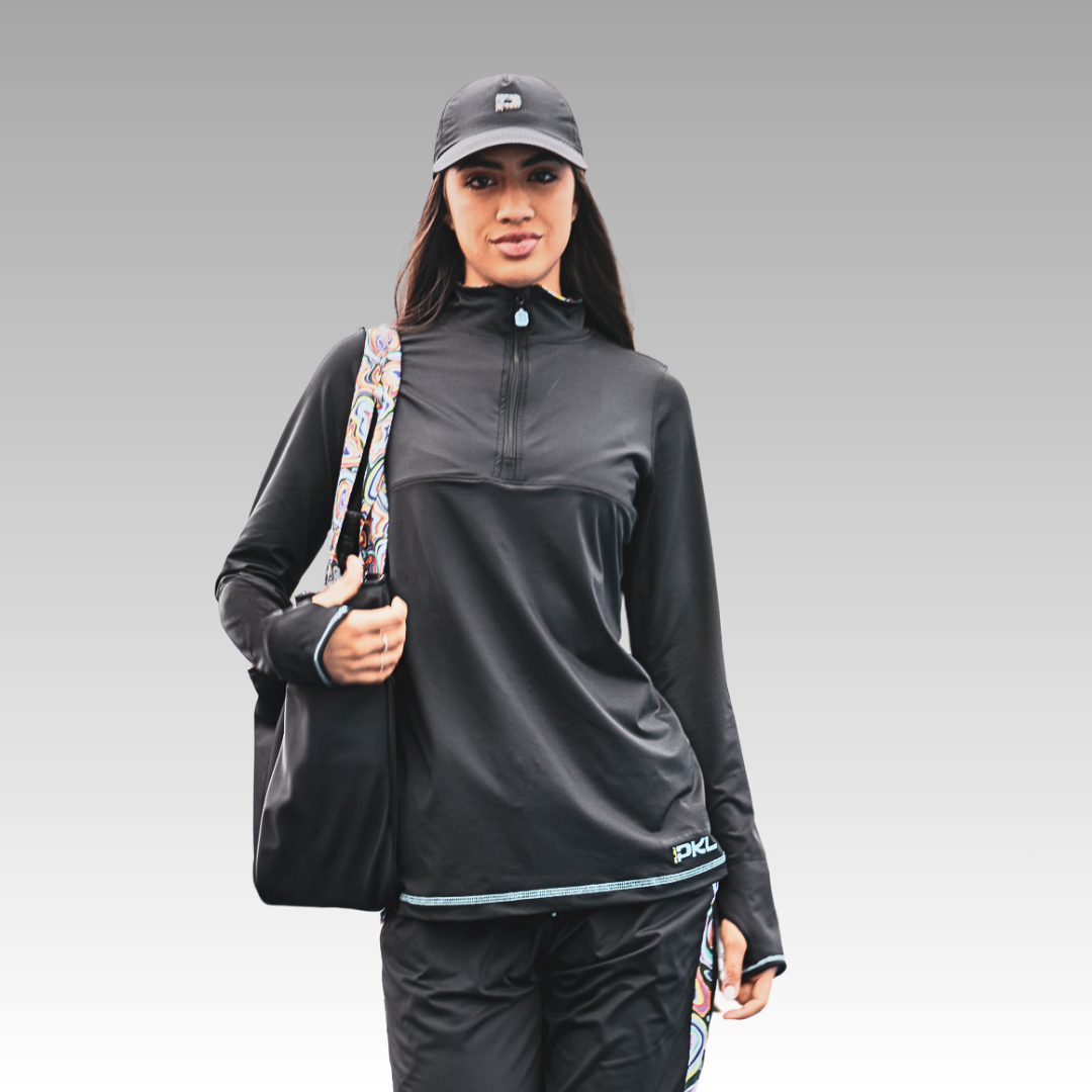 A woman models the Playly Store Soleil Tech Shirt, featuring long sleeves and UV protection. She pairs it with a black athletic cap, black pants, and a black bag with a colorful strap, standing against a neutral gradient background.