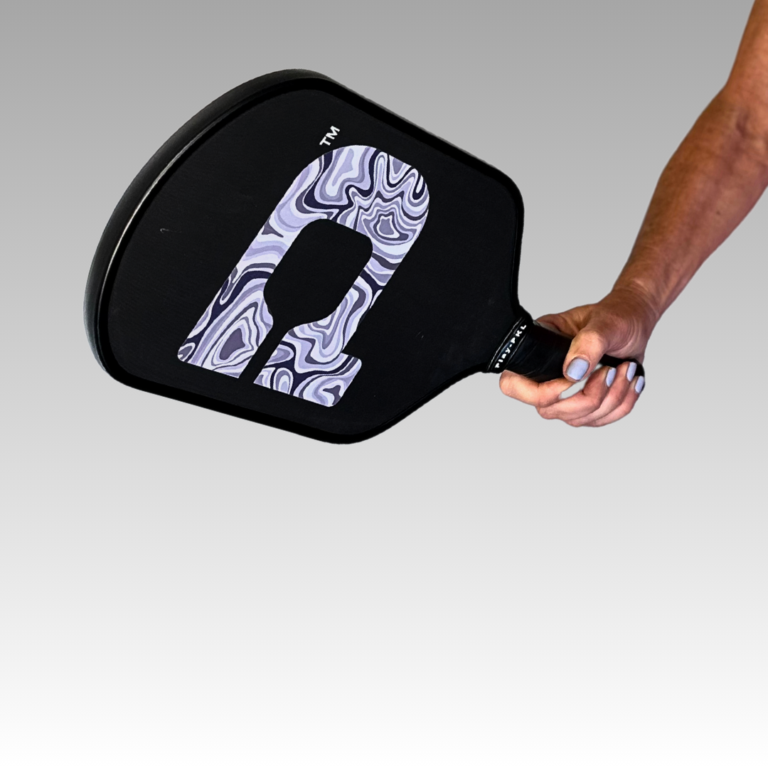 A right hand grips the Playly Store Signature Pro Pickleball Paddle, a black carbon fiber paddle with a large marbled “O” on its face. A smooth gray gradient forms the background, and part of the person’s arm is visible. USA Pickleball Approved.