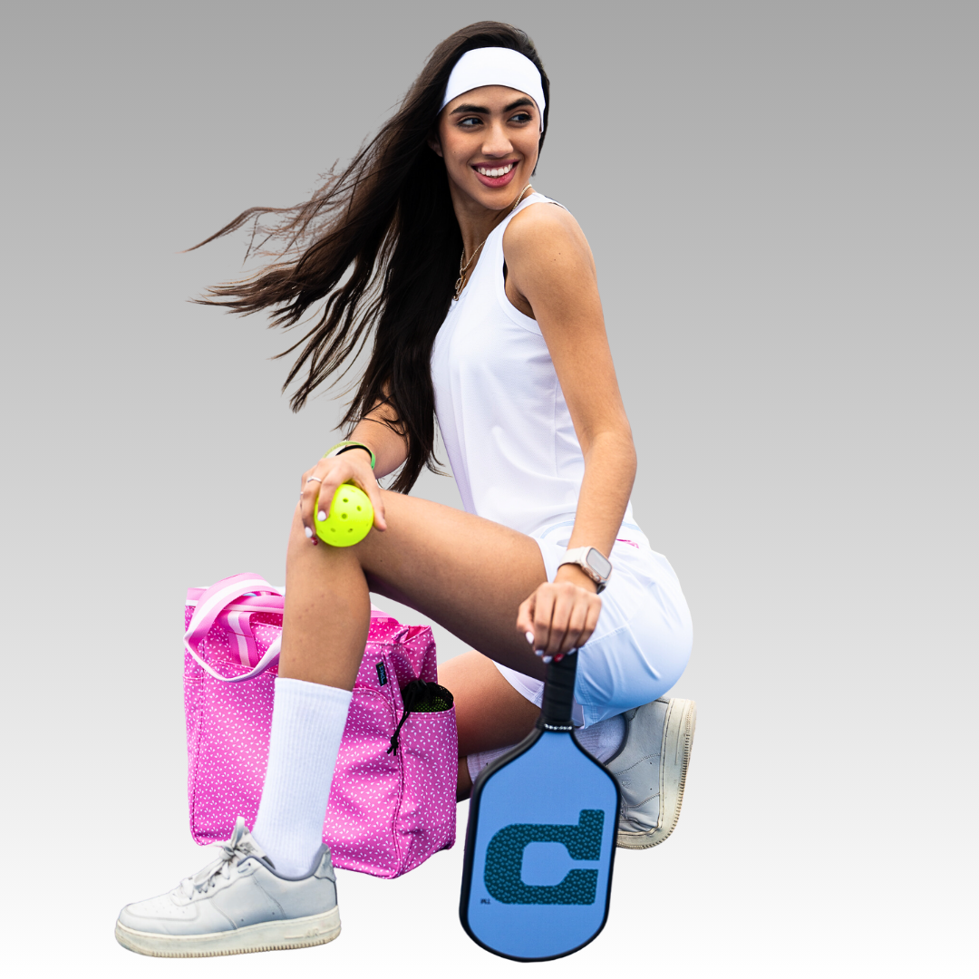 A smiling woman in a white athletic outfit and headband kneels with the Playly Store Signature Pro Pickleball Paddle, USA Pickleball Approved, holding a yellow ball next to a pink tote bag against a plain light gray background.