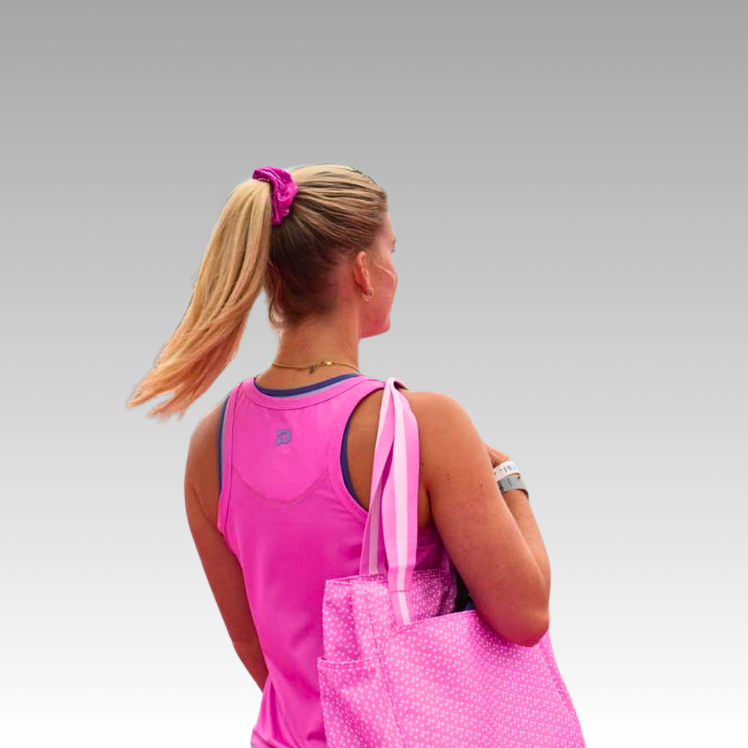 A woman with blonde hair in a ponytail, held by the Playly Store Shanti Pickleball Hair Scrunchie, wears a pink athletic tank top and carries a pink polka-dot tote, set against a gray-to-white gradient background.