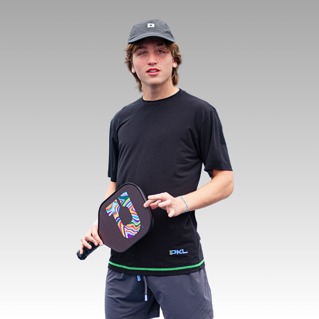 A young man in a Playly Store Warrior Tee, gray shorts, and a dark cap holds a colorful pickleball paddle. He stands against a light gray gradient background, looking at the camera with a relaxed expression.