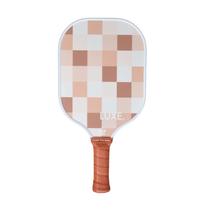 The LUXE Pickleball Checker paddle has a light brown grip band and a textured face with pink, tan, and beige squares in a grid. "LUXE." is printed near the bottom of the paddle, set against a plain white background.