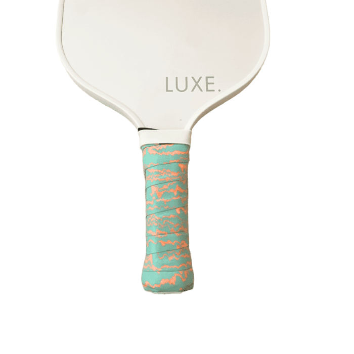 A white LUXE Pickleball paddle is shown with "LUXE." near its base and a teal Pickleball Paddle Grip featuring a wavy orange pattern, highlighting the unique style from LUXE Pickleball against a plain white background.