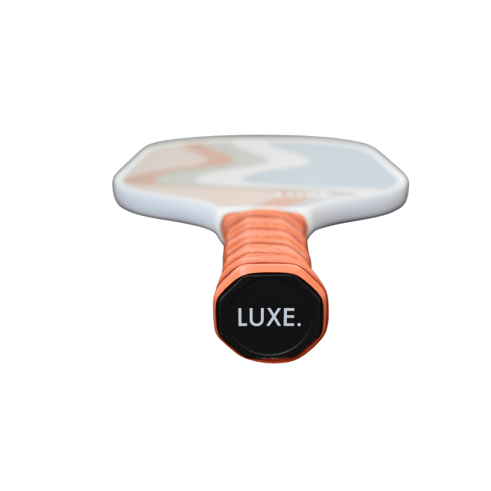 Close-up of The Luka paddle handle wrapped in orange grip tape, LUXE Pickleball in white on the black end cap. The blurred face shows a soft, wavy pastel pattern—perfect to elevate your game against a plain white background.