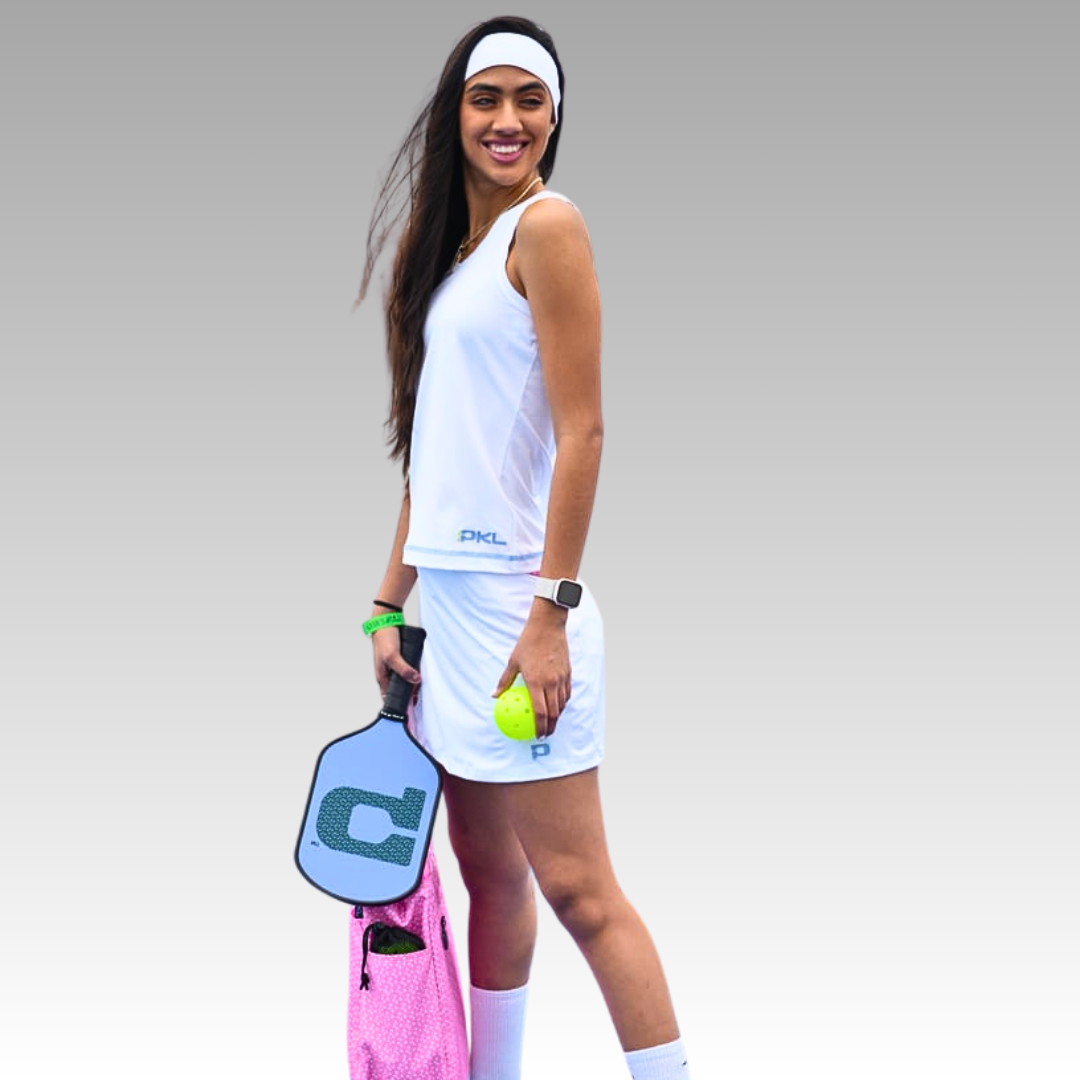 A woman in white athletic wear and headband smiles, holding a pickleball paddle and ball. She wears the Playly Store Sammy Skort 2.0 in CoolFlow fabric, carries a pink sports bag, and has a smartwatch against a plain gray background.