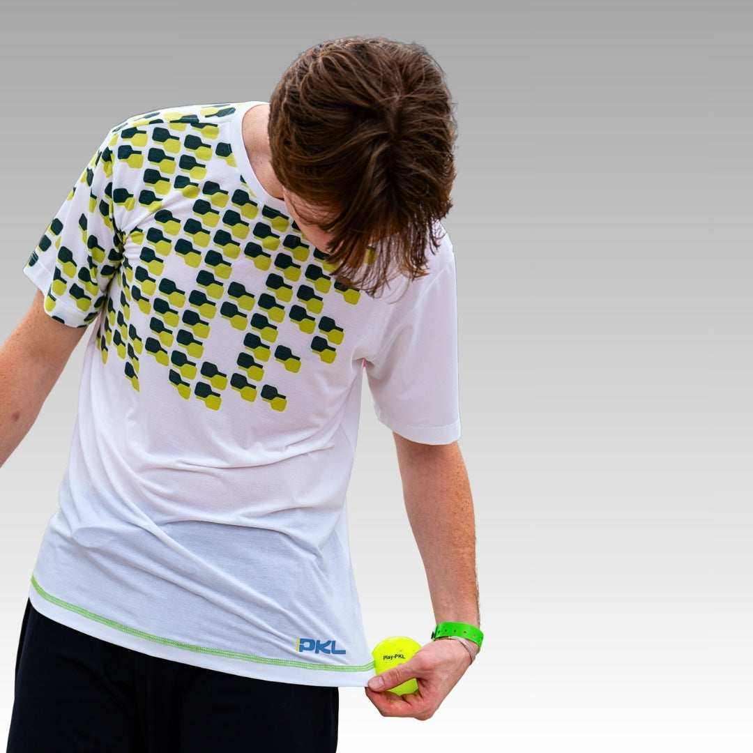 Wearing the Playly Store Warrior Tee, a moisture-wicking shirt with a green and black geometric pattern, a person looks down holding a yellow pickleball and pulling the hem, set against a smooth gray gradient background.
