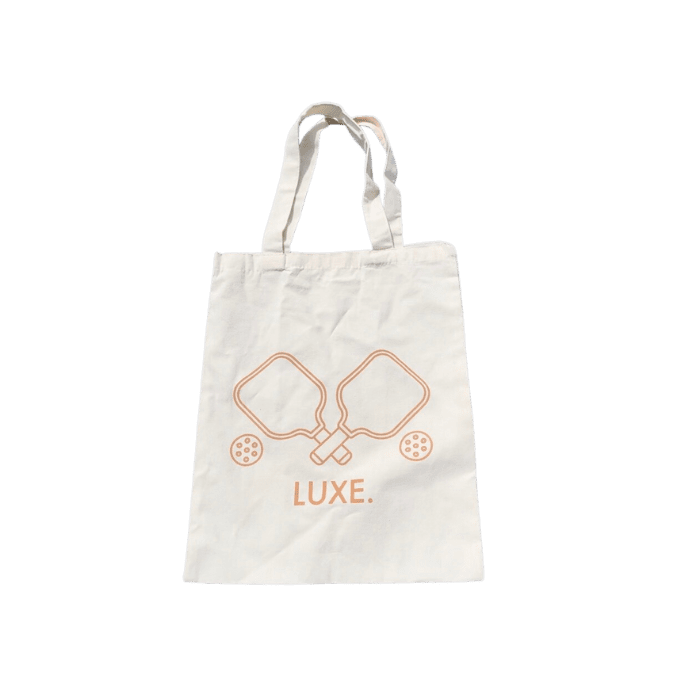 The LUXE Pickleball Tote Bag is a white bag with two long handles. The front features an orange line drawing of crossed paddles and balls above “LUXE.” Ideal for carrying pickleball gear, it’s shown flat on a plain white background.