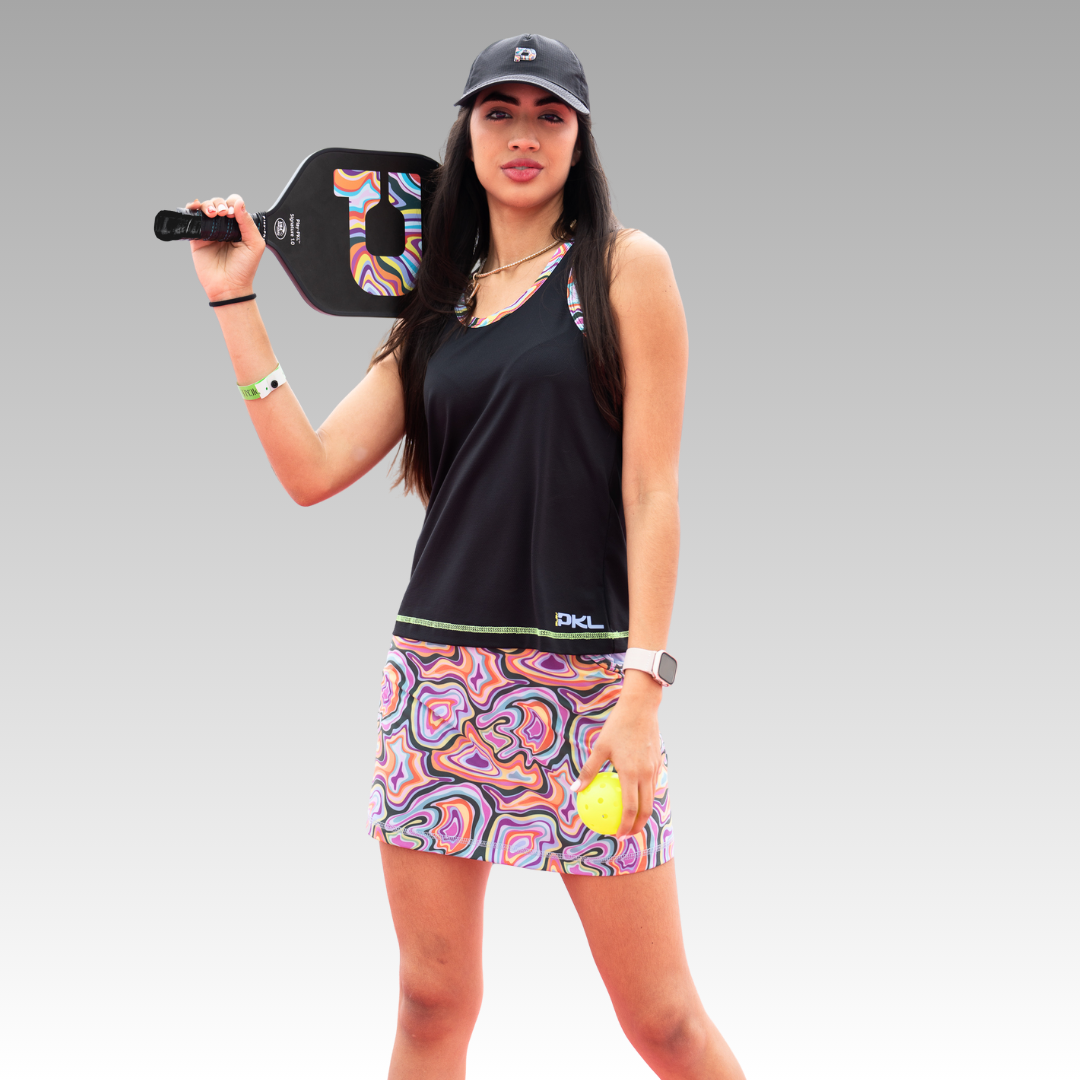A woman holds the Playly Store Signature Pickleball Paddle with a graphite surface over her shoulder and a yellow ball in her hand. She wears a black tank top, colorful skirt, black cap, smartwatch, and stands before a gray gradient background.