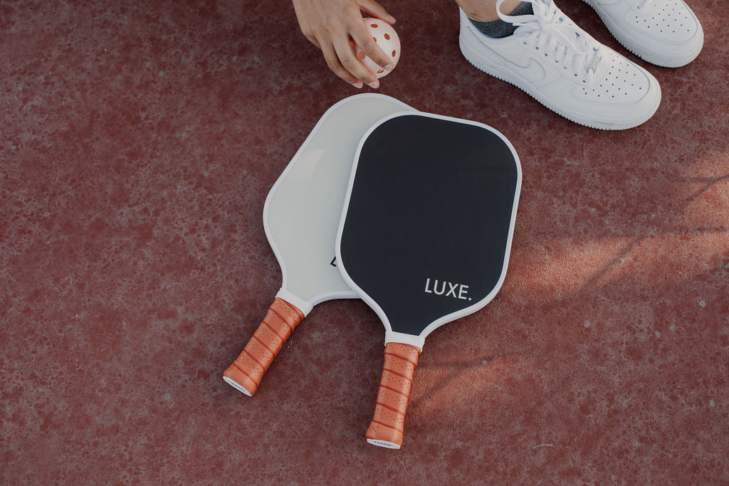 A person in white sneakers holds a white perforated ball near two LUXE Pickleball Aesthetic Solids - Ace paddles, one white and one black with brown handles, lying on a red textured court.