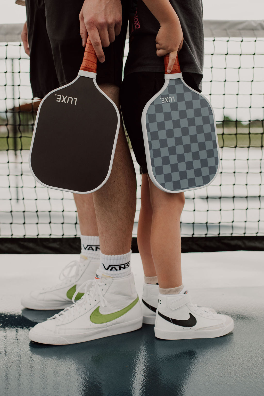 Two people, likely an adult and a child, stand on a glossy USA Pickleball court holding LUXE Pickleball Aesthetic Solids - Ace paddles. Both wear white Nike sneakers with black or dark shorts; a net is visible in the background.