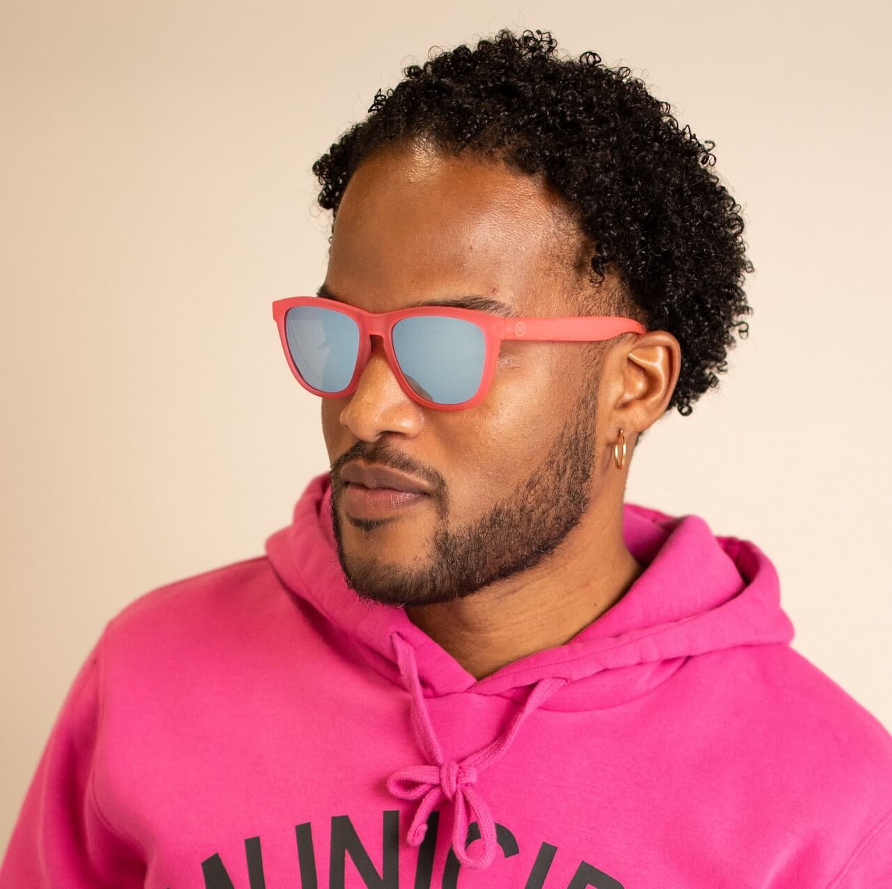 A man with short curly hair and a trimmed beard wears Epoch Eyewear VIBE sunglasses with reflective pink lenses and a bright pink hoodie. He sports a gold hoop earring, faces slightly left, and poses against a plain light background.