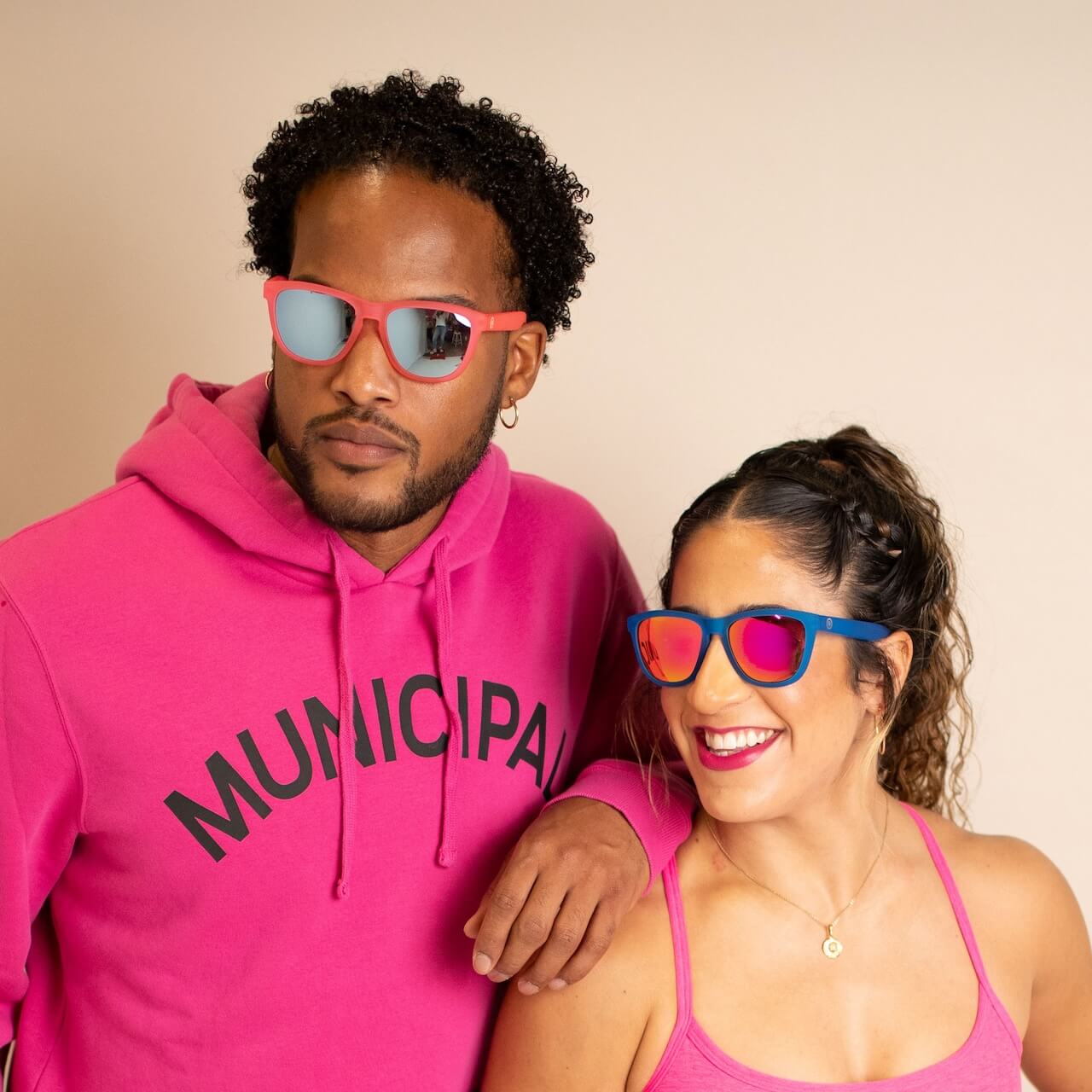 Two people wear vibrant Epoch Eyewear VIBE sunglasses and bright pink outfits against a beige background. The man has on a MUNICIPAL hoodie and coral VIBE shades, while the woman smiles in a pink tank top with blue polarized VIBE sunglasses.