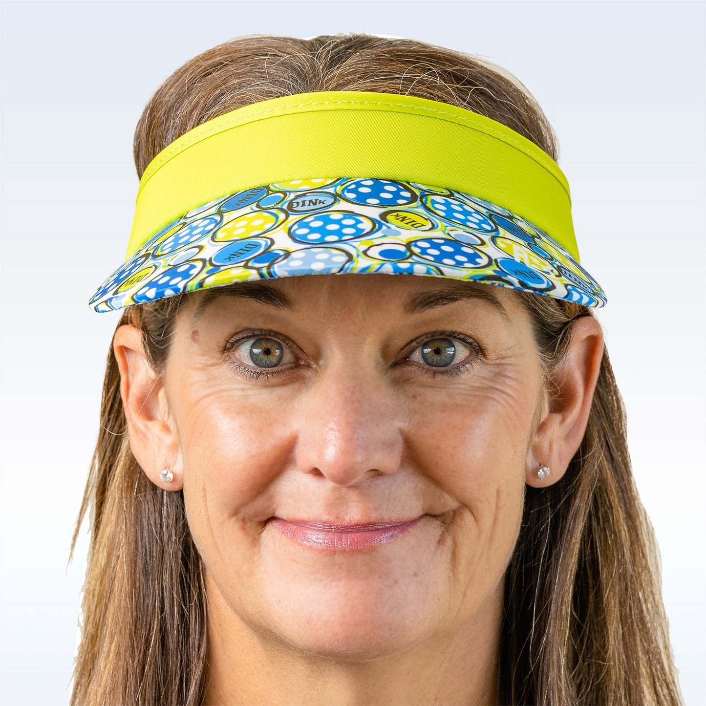 A middle-aged woman with light brown hair wears the Pickleball Bella PB Visor Dink 1, showcasing its blue, white, and yellow design. The lightweight, quick-dry visor pairs with her small stud earrings and faint smile against a plain backdrop.