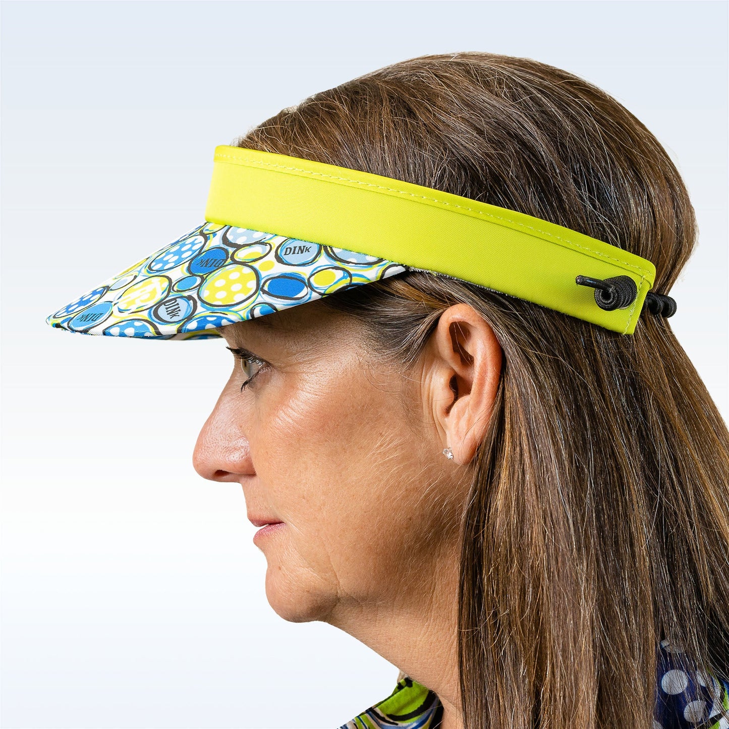 A woman with straight brown hair models the Pickleball Bella PB Visor Dink 1—a lightweight, quick-dry visor with a yellow band, blue/green/white circle brim, adjustable cord back—shown in profile against a light background.