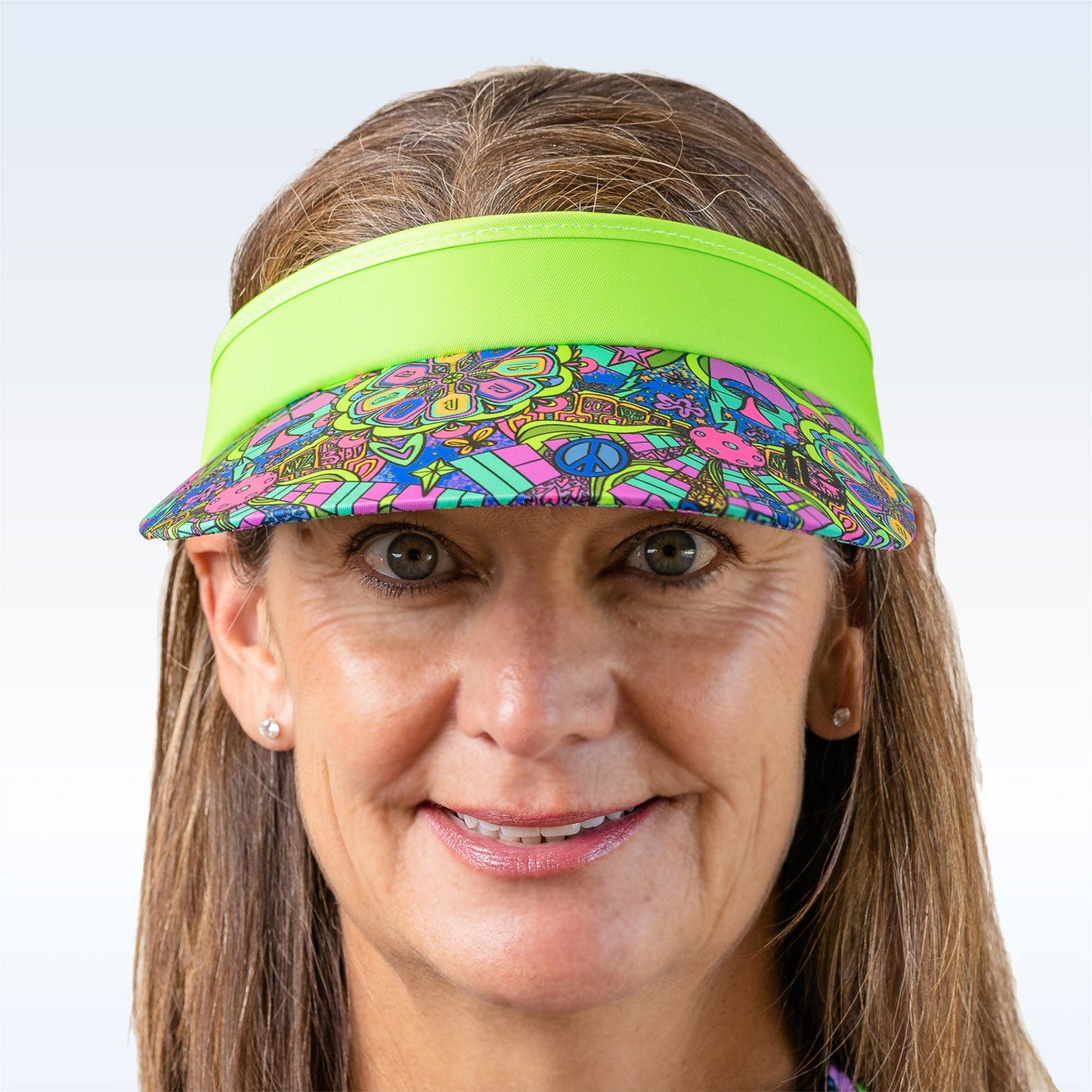 A smiling middle-aged woman with long brown hair wears the PB Visor Groovy by Pickleball Bella, featuring peace signs, flowers, and butterflies. This lightweight, quick-dry accessory is paired with stud earrings on a pale blue background.