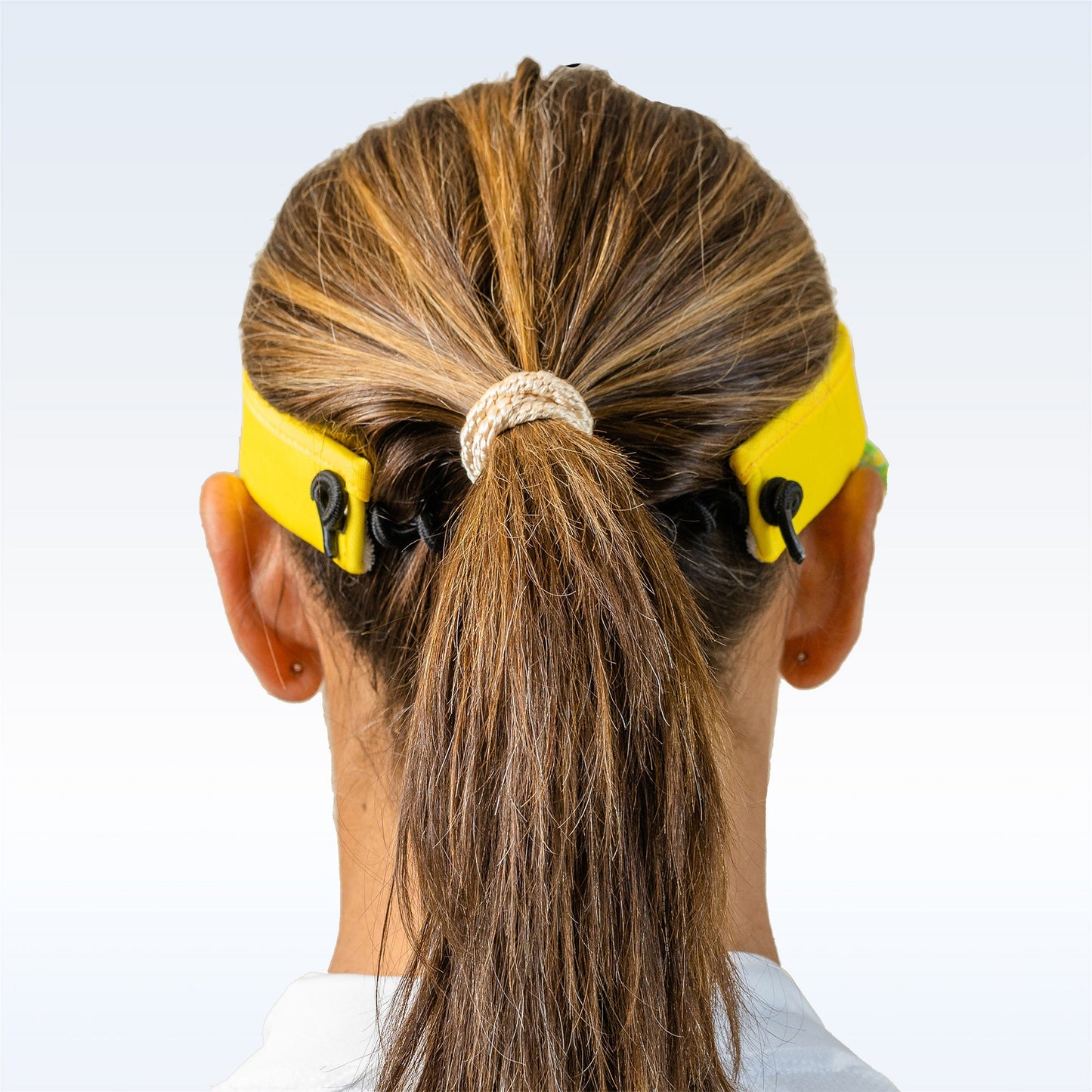 A woman with brown hair in a ponytail, secured by a light hair tie, is shown from behind wearing the Pickleball Bella PB Visor Palms 1—a lightweight, quick-dry yellow strap connecting her face mask straps behind her head.