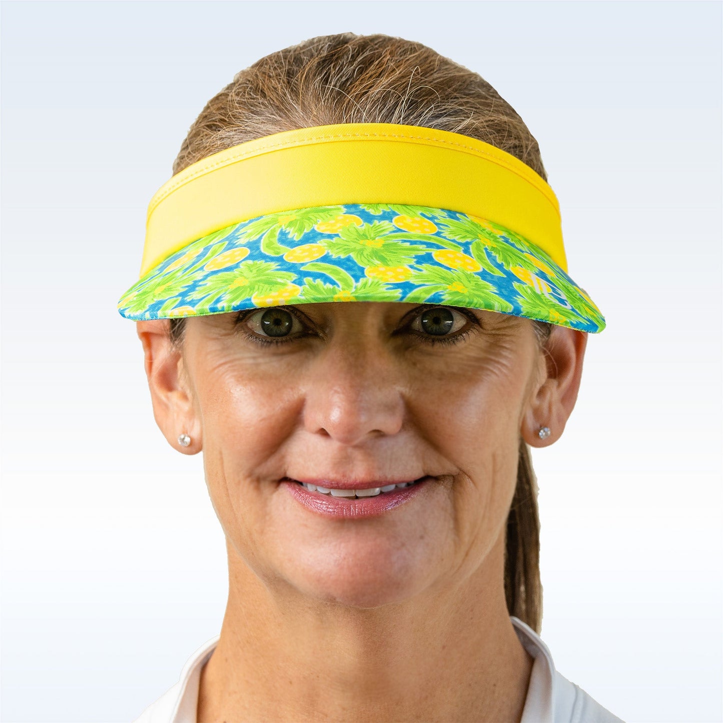 A smiling woman with light brown hair tied back wears the Pickleball Bella PB Visor Palms 1, featuring a yellow and green palms print, along with a white shirt and small stud earrings. The background fades from pale blue to white.