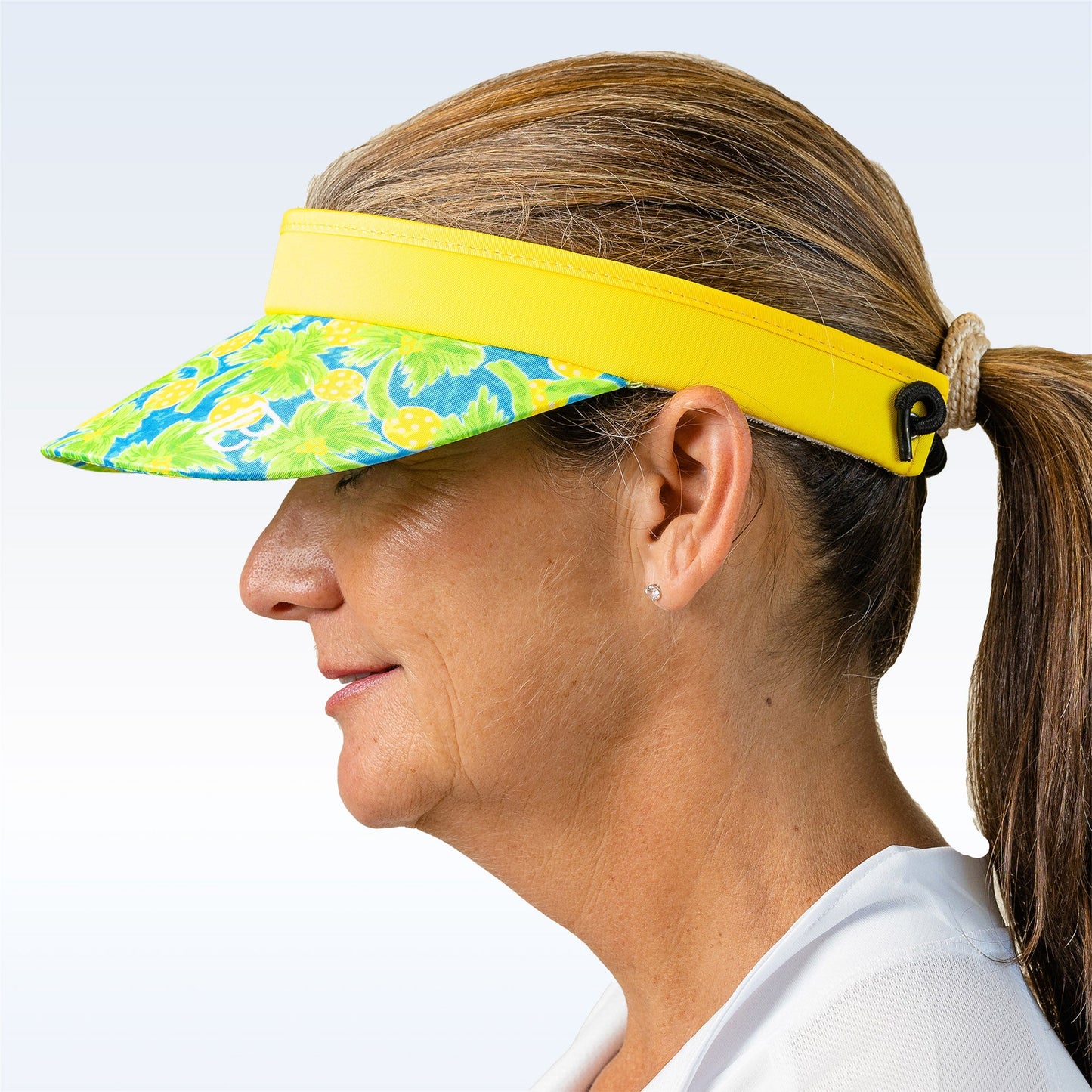 A woman with a low ponytail wears the Pickleball Bella PB Visor Palms 1—featuring a vibrant yellow and blue palms print—paired with stud earrings and a white top, set against a light gradient background for a fresh, clean look.