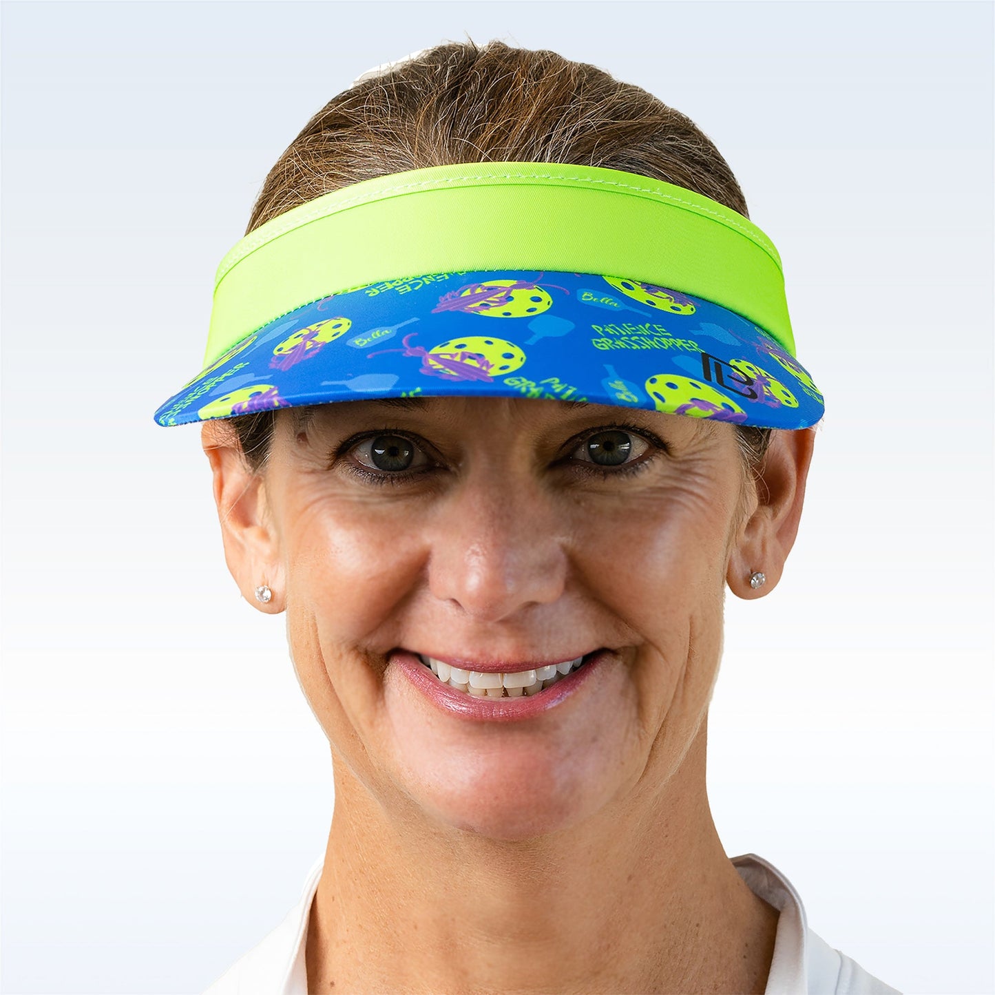 A smiling woman with light skin and brown hair wears the neon green and blue Pickleball Bella PB Visor Patience Grasshopper, holding yellow paddles and balls. She pairs stud earrings with a light collared shirt against a neutral background.