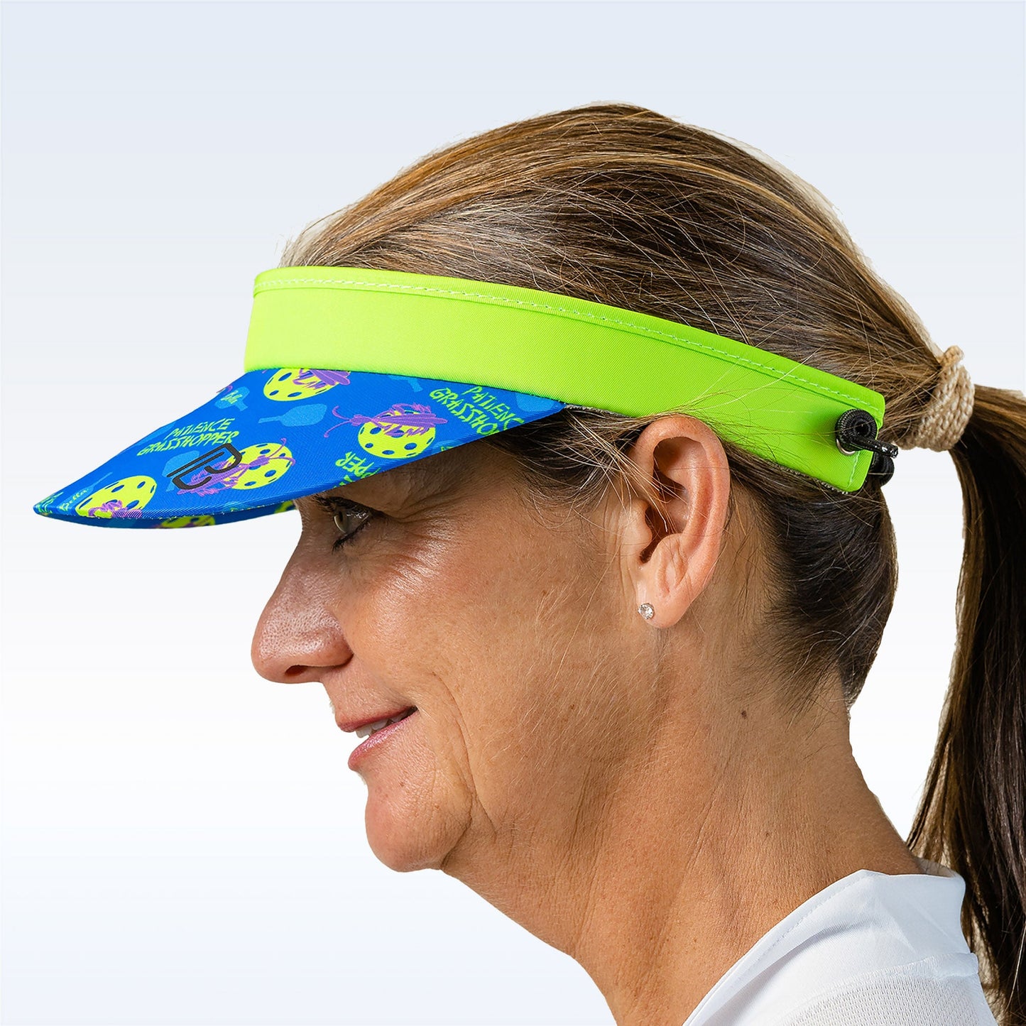 A smiling woman in profile wears the Pickleball Bella PB Visor Patience Grasshopper with playful yellow cat faces and vibrant designs. Her brown hair is in a low ponytail, and she sports a white shirt against a soft gradient background.