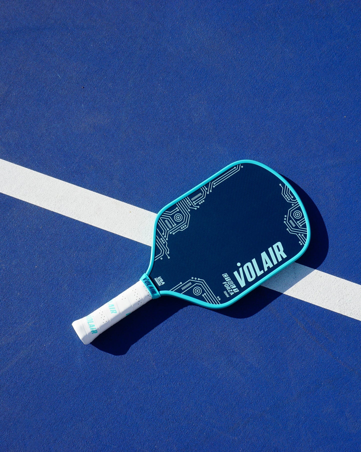 A Volair Pickleball CTRL 1 16mm Forza paddle with a blue and black circuit board design rests on a blue court, crossing a white sideline. The handle features white grip tape and the scene is brightly lit by sunlight.