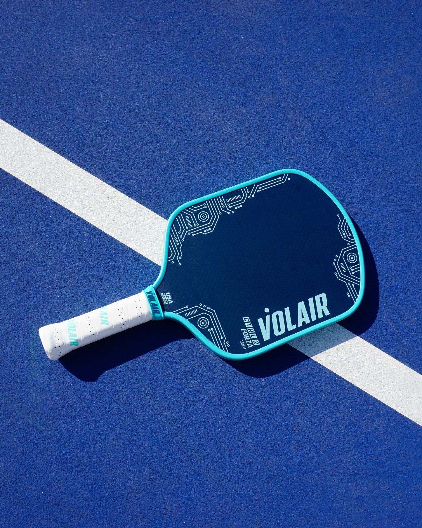 A blue Volair Pickleball CTRL 2 Forza 16mm paddle with circuit-like patterns and a white handle rests on a blue court, partly over a white line. The paddle features the Volair logo in bold white letters on its face.