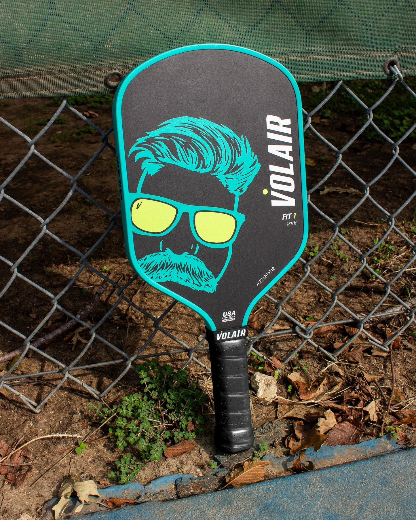 The Volair Pickleball Fit 1 16mm Limited Edition Paddle, featuring a black and turquoise design with an illustrated face in sunglasses, hair, and mustache plus vertical VOLAIR branding, leans on a chain-link fence near dry leaves on a dirt court.