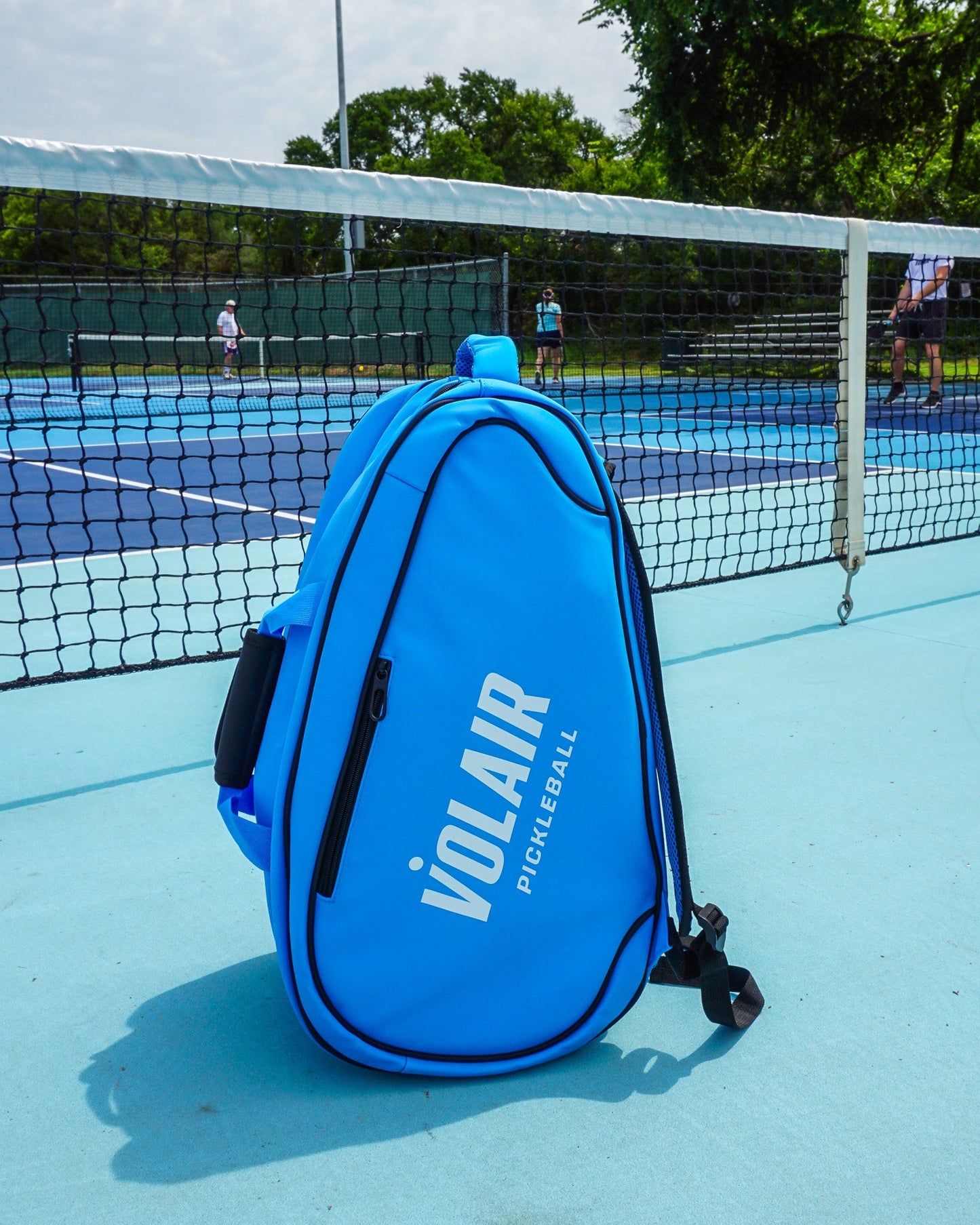 A Volair Pickleball Volair Paddle Bag in bright blue faux leather stands upright on a light blue court near the net, while people play pickleball on nearby courts surrounded by greenery and a partly cloudy sky.