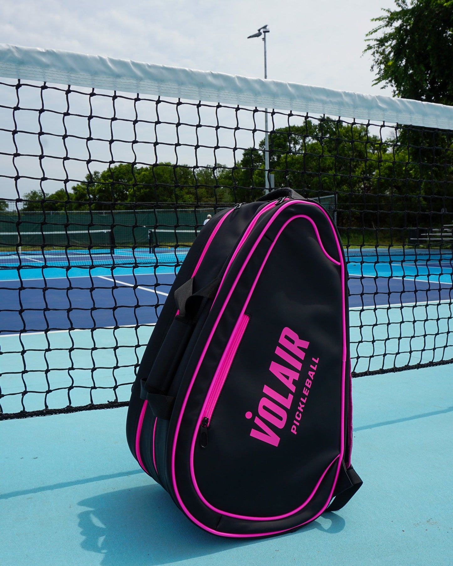 The Volair Paddle Bag by Volair Pickleball, featuring black faux leather with bright pink accents, leans against a net on an outdoor blue court with green trees and fences in the background under a clear sky.