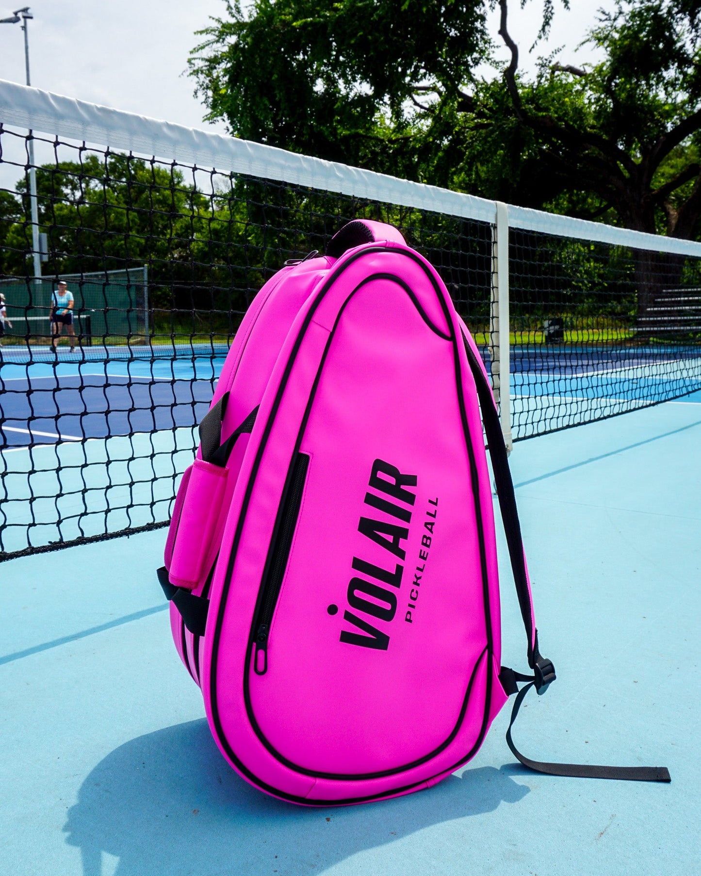 The Volair Paddle Bag by Volair Pickleball, in bright pink faux leather with black zippers and accents, stands upright on a blue pickleball court near the net, with sunlit trees and greenery in the background.