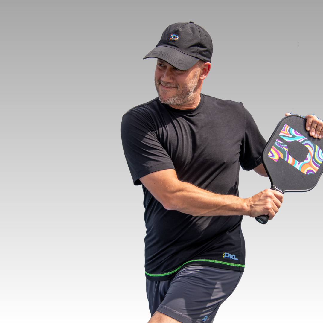 A man in a black Playly Store Warrior Tee and hat holds a colorful pickleball paddle, ready to swing. He smiles, wearing gray athletic shorts and an athletic fit moisture-wicking shirt against a light gray background.