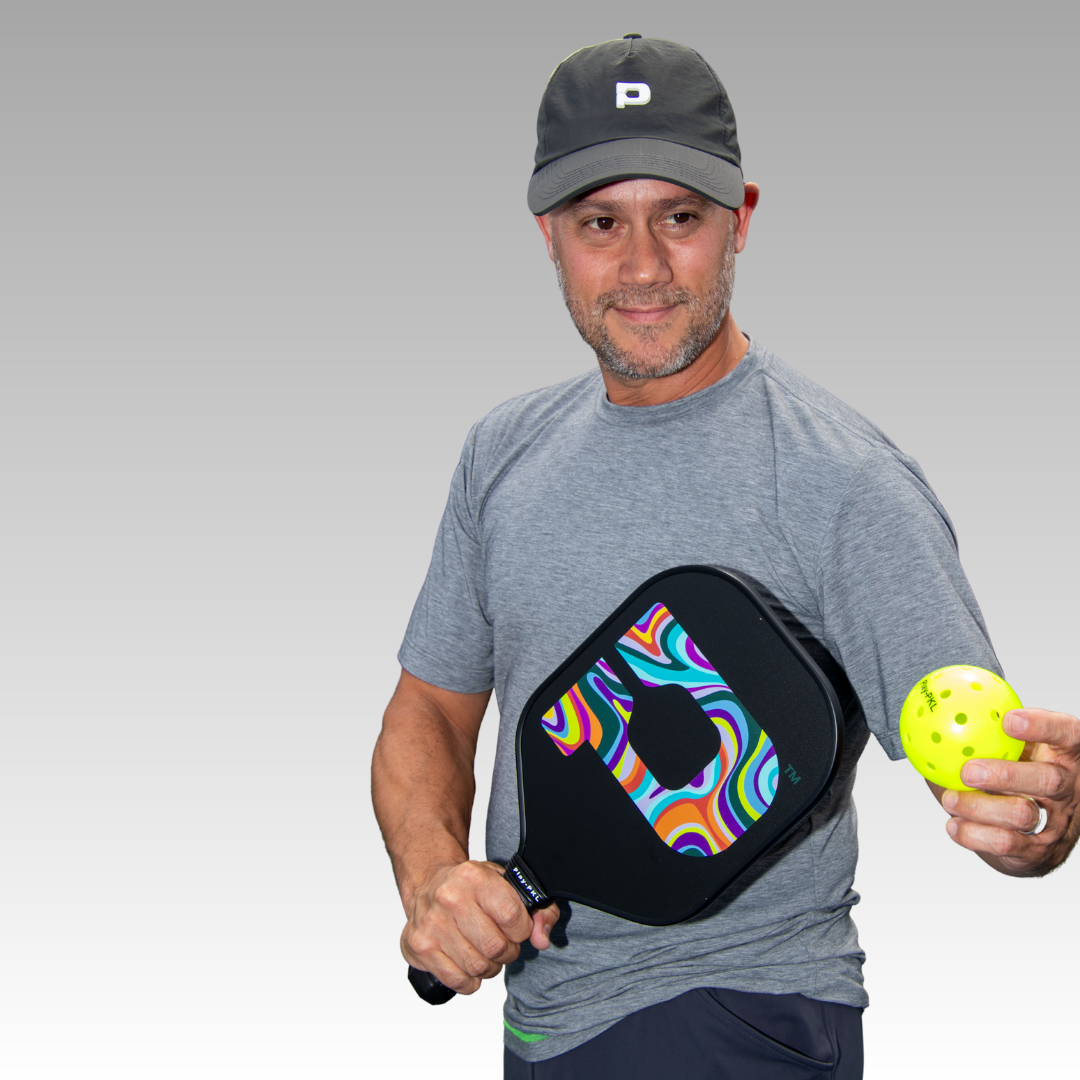 A man wearing the Playly Store Warrior Tee and matching cap holds a colorful pickleball paddle and a yellow pickleball, posing to serve. The background fades from gray to white.