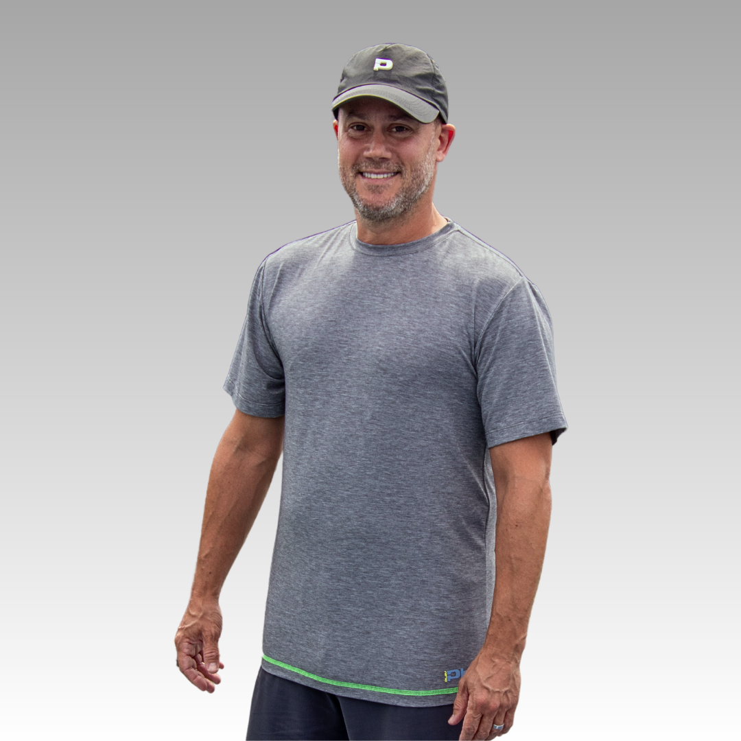 A smiling middle-aged man wears the Playly Store Warrior Tee, an athletic-fit gray pickleball shirt with green trim, plus a dark baseball cap, standing against a gradient gray background with his right arm relaxed at his side.