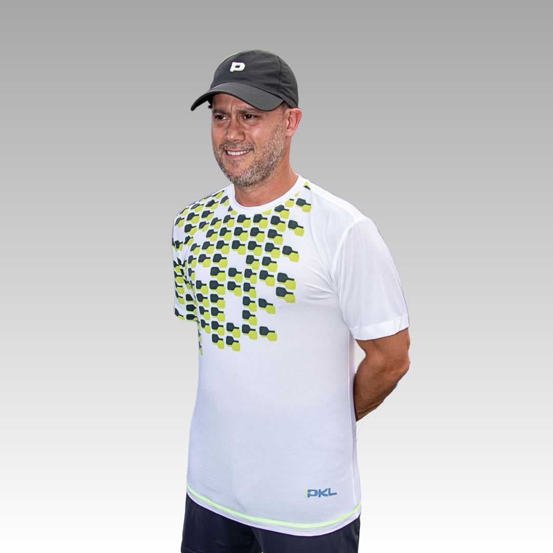 A man wearing Playly Store’s Warrior Tee—a white moisture-wicking shirt with a yellow and black geometric pattern on the upper left and "PKL" near the hem—stands smiling in a black baseball cap against a gray gradient background.