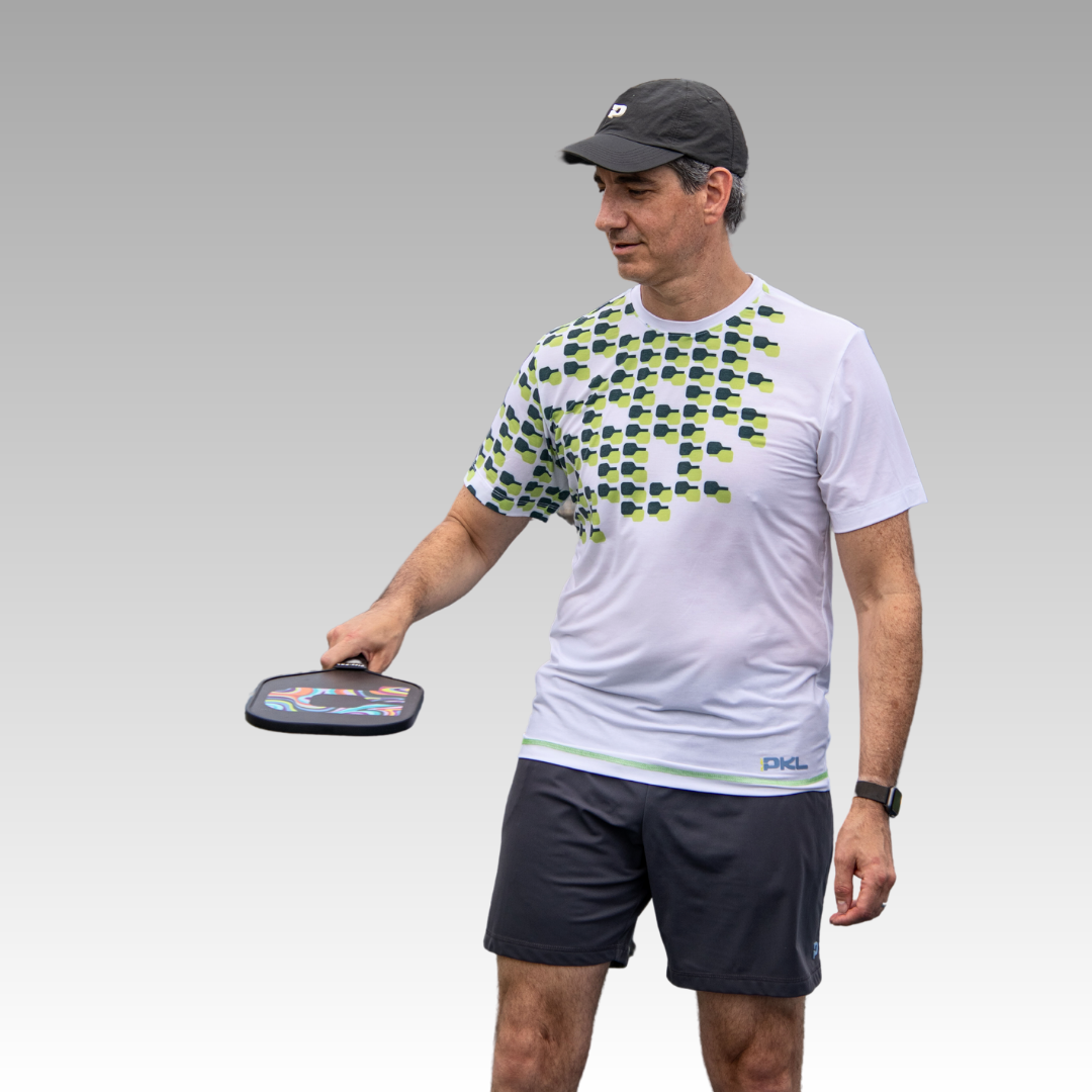 A man wearing the Playly Store Warrior Tee in an athletic fit, black shorts, black cap, and a black watch on his left wrist holds a paddle in his right hand, looking to his left with a relaxed posture against a plain grey background.