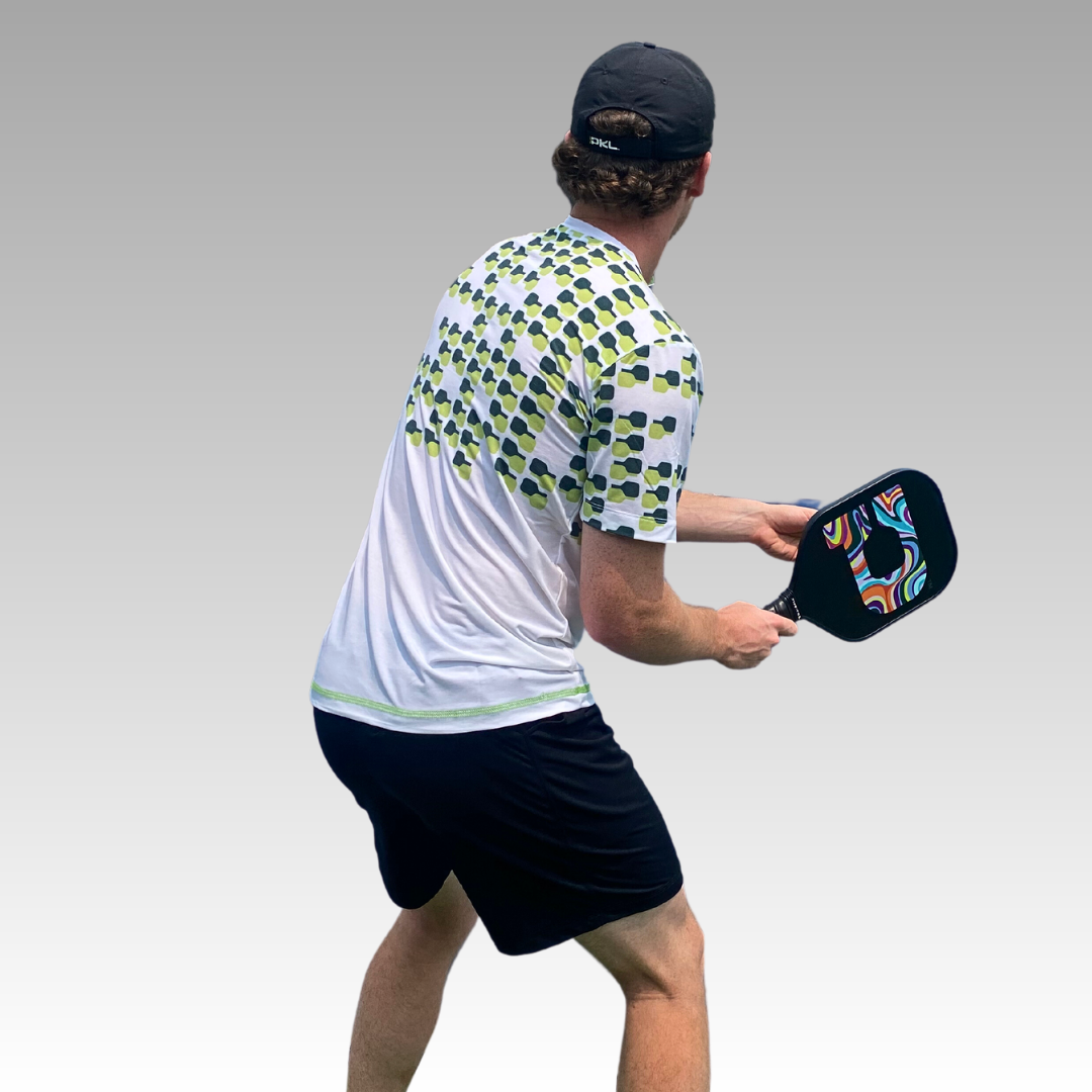 A man in a Playly Store Warrior Tee, black shorts, and a black cap stands ready to play pickleball, holding a colorful paddle and facing away from the camera against a gray gradient background.
