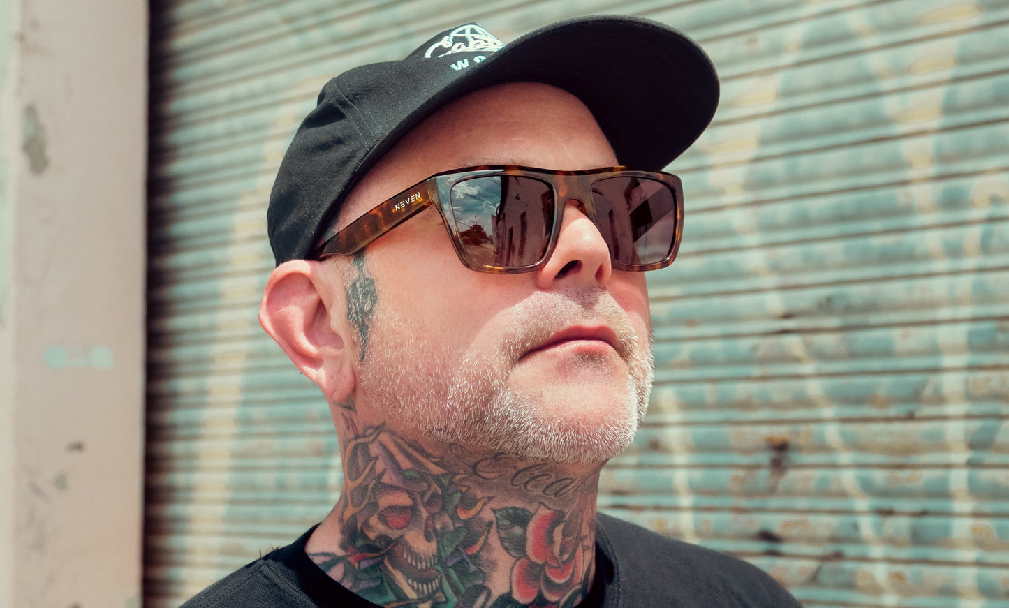A man wearing Neven Eyewear's Sepia polarized sunglasses and a black cap stands before a metal shutter. With a short gray beard and floral skull neck tattoos, he gazes upward in bright light, expression neutral.