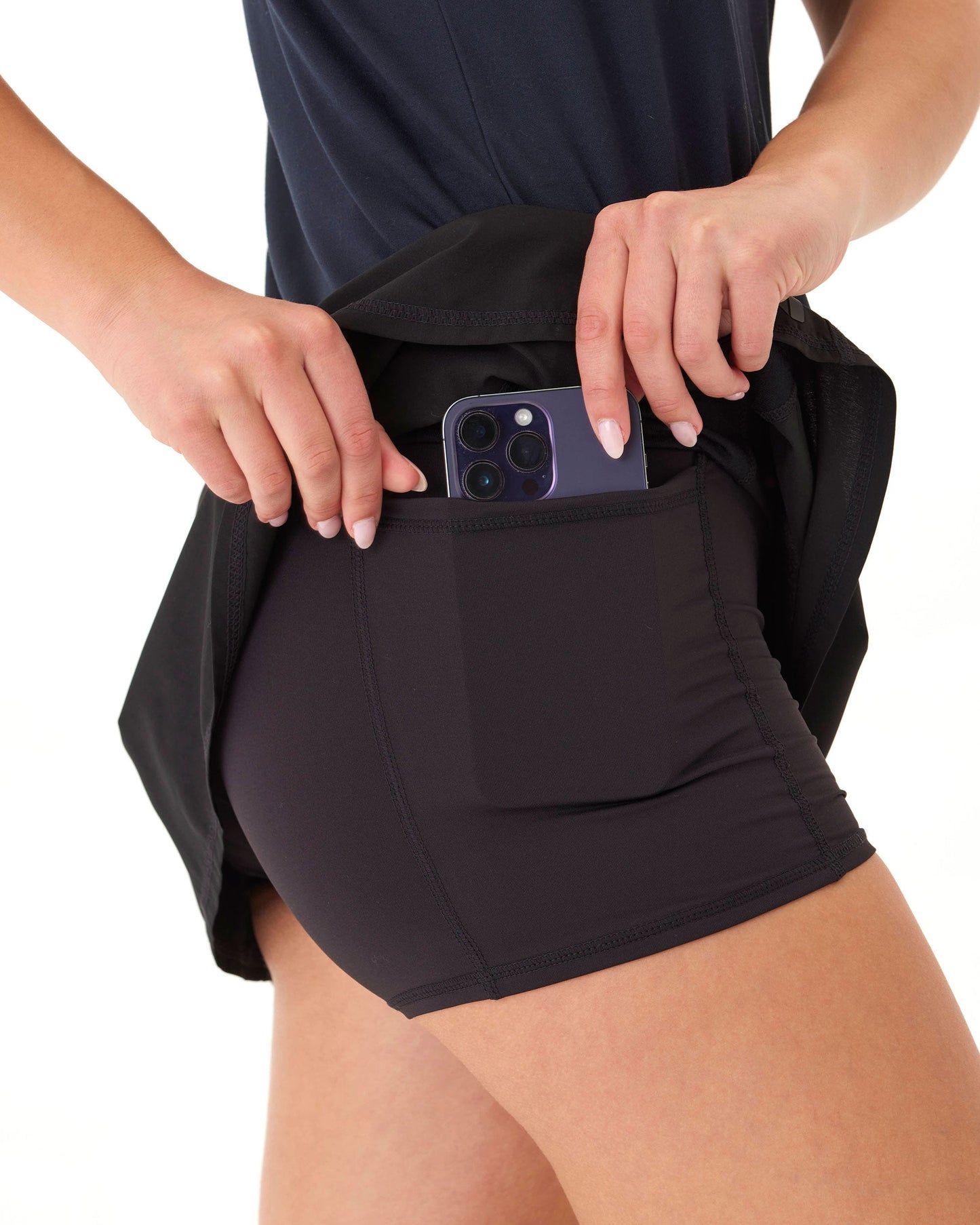 A person wearing a Wringer Wear Moab Skirt with a side pocket places a triple-lens smartphone into the pocket. Only the lower torso and hands are visible, dark shirt partly lifted, against a plain white background.