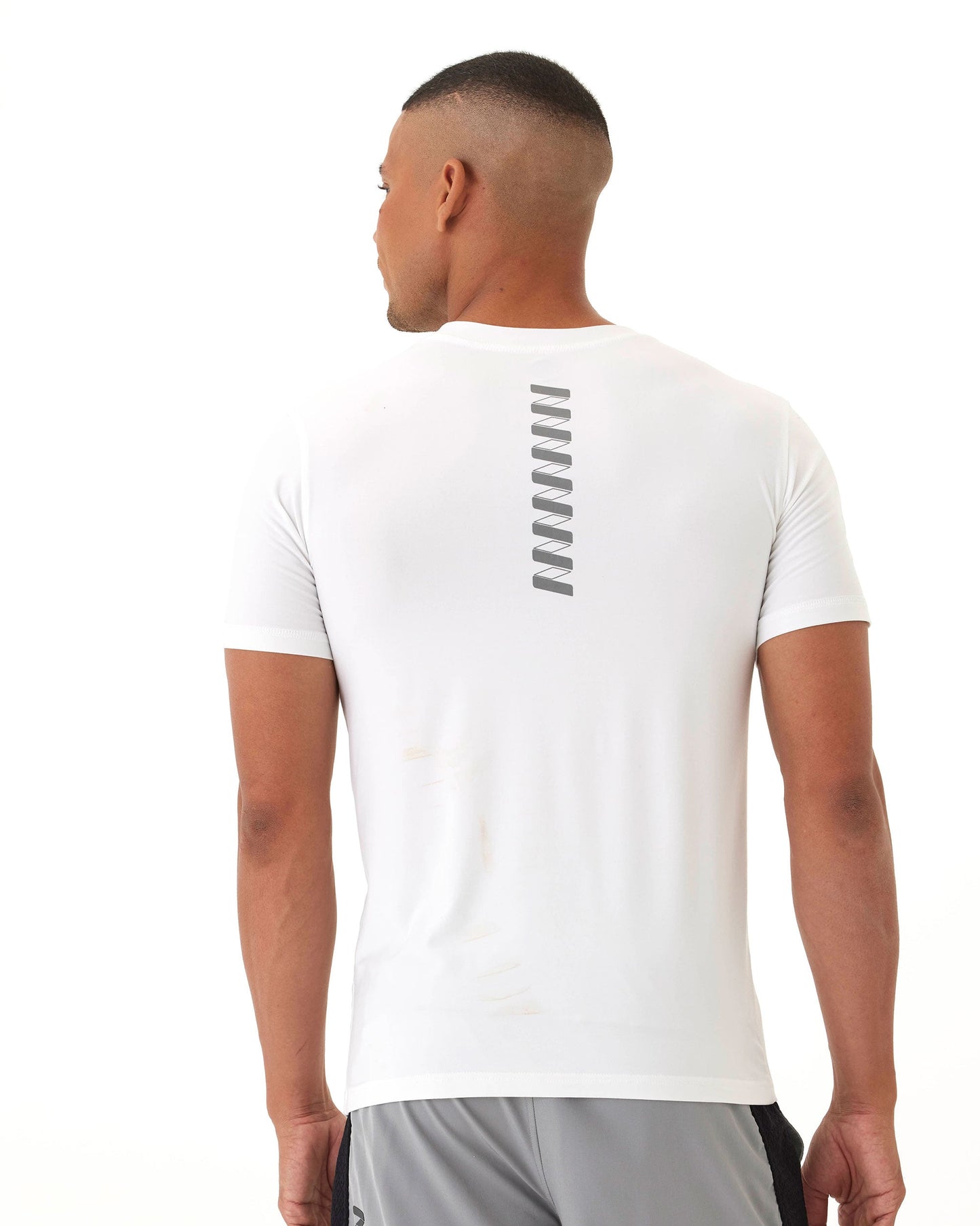 A man with short hair, seen from behind, wears the Wringer Wear Men's Short Sleeve Logo T-shirt featuring a vertical gray zigzag stripe on the upper back. He pairs it with light gray shorts against a plain white background in a relaxed pose.